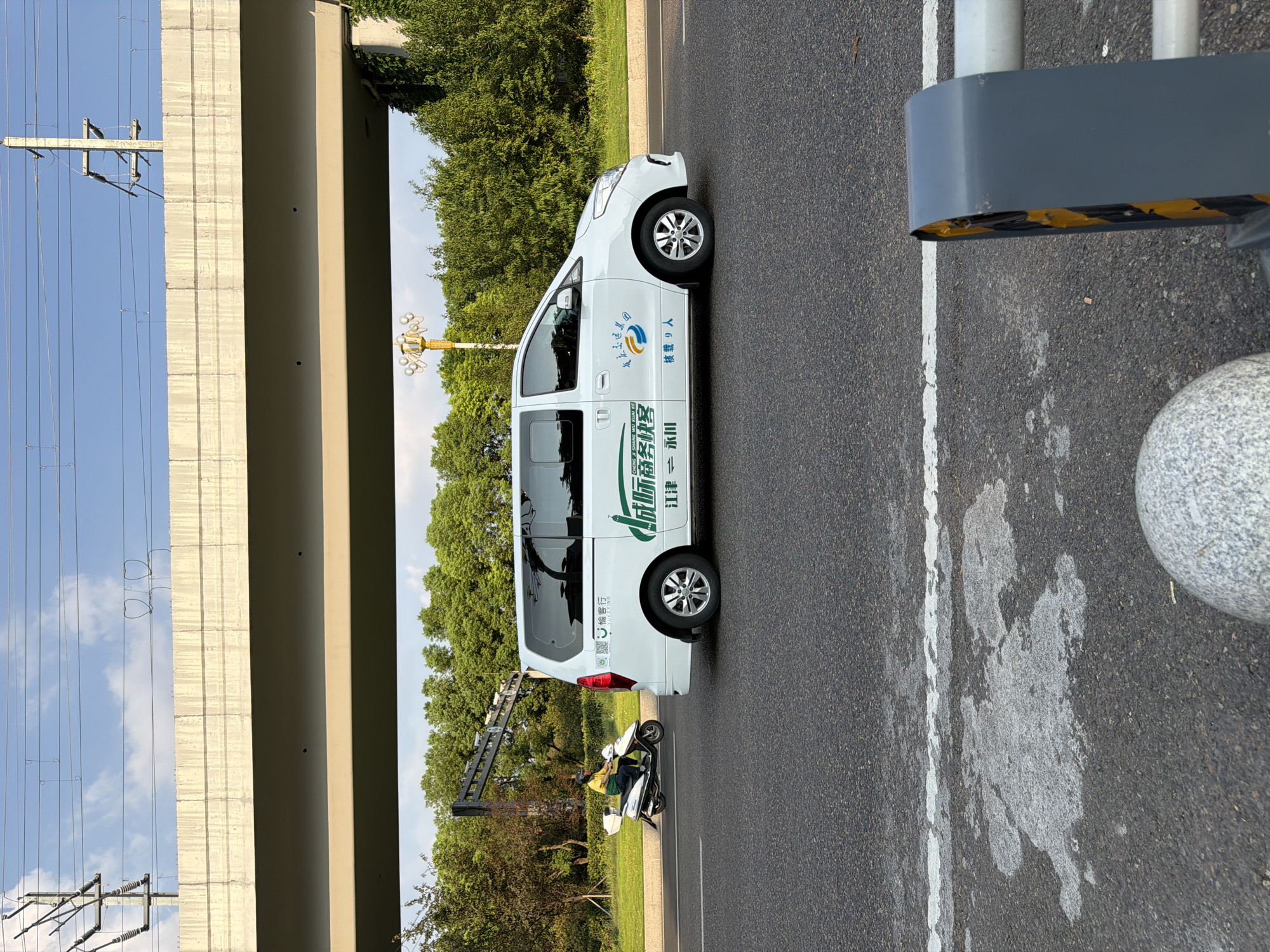 In the city of Jiangjin, China, a white passenger van with Chinese text and graphics indicating it's a "new business express" service is driving on an asphalt road under a concrete overpass. The van has the characters "江津" (Jiangjin) and "永川" (Yongchuan) on its side, suggesting a route or service area. A person on a white scooter is riding in the same direction ahead of the van. The environment is characterized by lush green trees and a clear blue sky with scattered clouds. Overhead, power lines stretch across the sky, supported by utility poles. The foreground of the image shows a portion of the road with a white line and some weathered patches, as well as a grey roadside barrier and a rounded granite bollard.