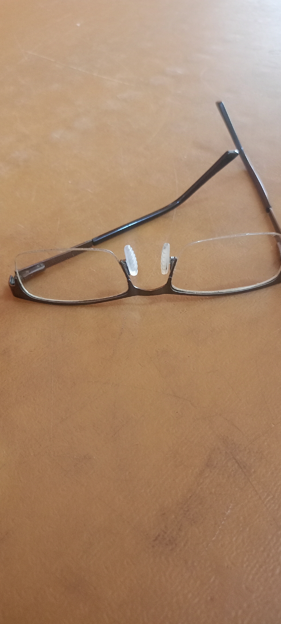 The image shows a pair of broken eyeglasses lying on a brown, textured surface. The eyeglasses appear to be half-rimmed, with clear lenses and thin dark frames. One of the temples is bent upwards at an angle, indicating damage. The surface resembles a wooden table with visible grain and some minor scuff marks. The lighting suggests an indoor environment. No people are visible in the image. The image is taken from a slightly elevated angle, looking down at the eyeglasses. The location in the city of Mekar Rahayu, Indonesia, is not depicted or suggested by any elements within the photograph.