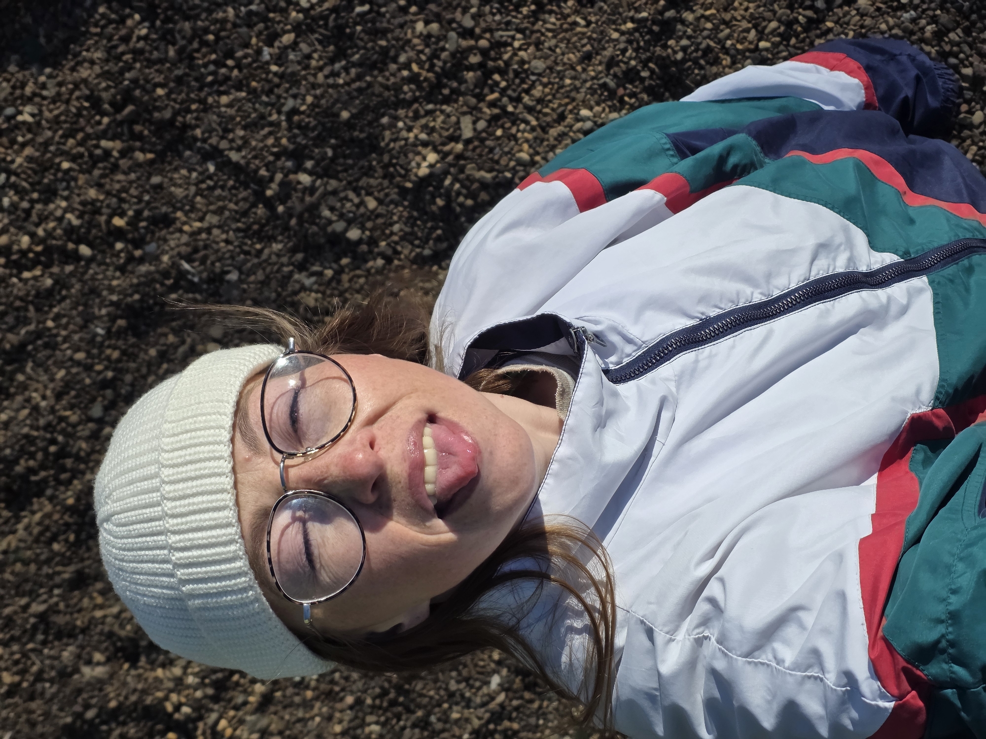 A person lies on what appears to be a gravel surface. They are wearing a white beanie and round glasses, and their tongue is sticking out. They are also wearing a white jacket with green, red, and possibly navy blue accents. The gravel surface appears to be composed of small, dark stones. The photo is taken from above, with the person's face angled upward towards the camera. The location is in Chaikovsky, Russia.