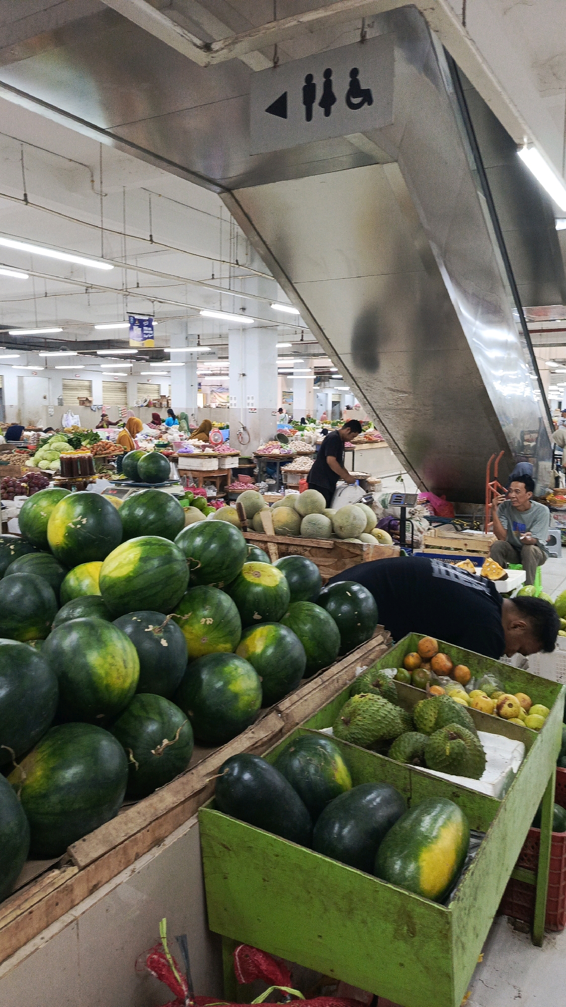 The image depicts a bustling indoor market in Nologaten, Indonesia, likely a public market given the layout and the presence of a large escalator. The foreground is dominated by a display of fresh produce, primarily watermelons in wooden crates. To the right, another green wooden crate contains various fruits, including what appear to be soursop and possibly papayas or other yellow-skinned fruits.
In the mid-ground, several people are engaged in market activities. A man in a black t-shirt is tending to produce. Further in the background, a broader view of the market reveals numerous stalls laden with a variety of fruits and vegetables, with many people browsing and shopping. The market interior is well-lit by overhead fluorescent lights, and the architecture suggests a large, covered space. Above the escalator, a directional sign points to restrooms, indicated by restroom symbols for men, women, and a wheelchair-accessible facility. The overall atmosphere is one of commerce and daily life in a busy Indonesian market.