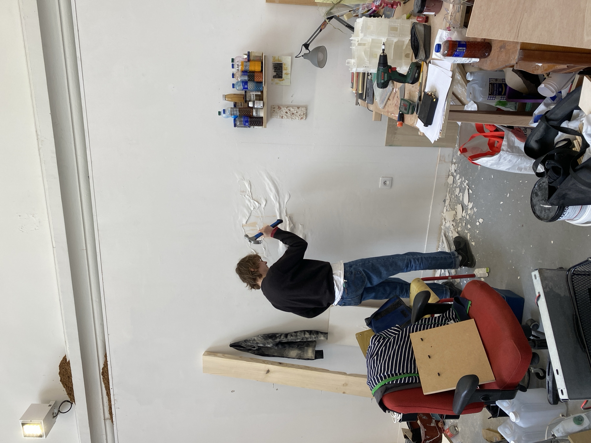 A person is peeling wallpaper from a white wall. The person has light brown hair, is wearing a black sweatshirt, blue jeans, and black shoes, and is holding a hammer. The room is cluttered, with a desk covered in objects, a red office chair, and debris on the floor. There is a shelf with bottles and jars on the wall. There's an electrical outlet on the wall. A wooden board is leaning against the wall. It is not possible to confirm the exact location within Paris, France based on the image.