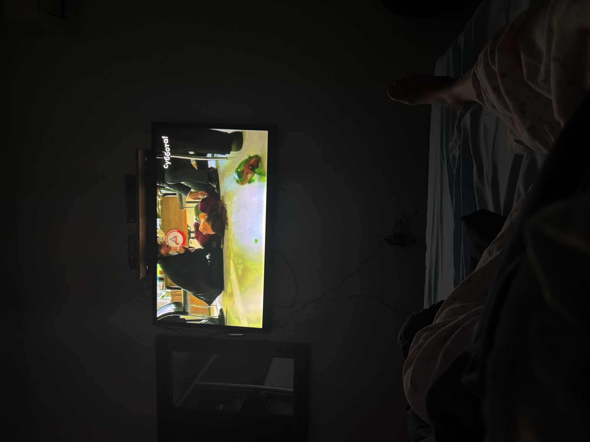 The image depicts a dimly lit bedroom scene, likely at night, with the main light source emanating from a flat-screen television. The photo is taken from a slightly elevated perspective, looking towards a bed and a wall-mounted TV.
On the right side of the frame, a person's bare foot and lower leg are visible, resting on a bed covered with a light blue sheet. The person appears to be wearing light-colored, patterned sleepwear, with a darker blanket or clothing partially obscuring their body.
The central focus is a wall-mounted flat-screen television displaying a vibrant scene. The screen shows two individuals in an indoor setting. One person, with dark hair, is seen from behind, wearing a dark top and leaning over a surface, possibly interacting with an object. A bright green and reddish-brown object, resembling food or a toy, is visible on a surface in front of them. Prominently displayed vertically along the left edge of the TV screen content is the Cyrillic word "суббота!" (Subbota!), which translates to "Saturday!". Additionally, a small red circular logo containing the number "4" is visible in the bottom-left corner of the TV's content, likely indicating the channel.
Below the television, a dark, reflective piece of furniture, possibly a dresser or TV stand, is partially visible. Wires are seen trailing from the TV area down the wall. The overall mood conveyed is one of relaxation and comfort, with someone unwinding by watching television in bed. The time of day is clearly night due to the darkness of the room, with the television providing the primary illumination. While the location context is Budva, Montenegro, there are no specific visual cues within the image to identify this location.