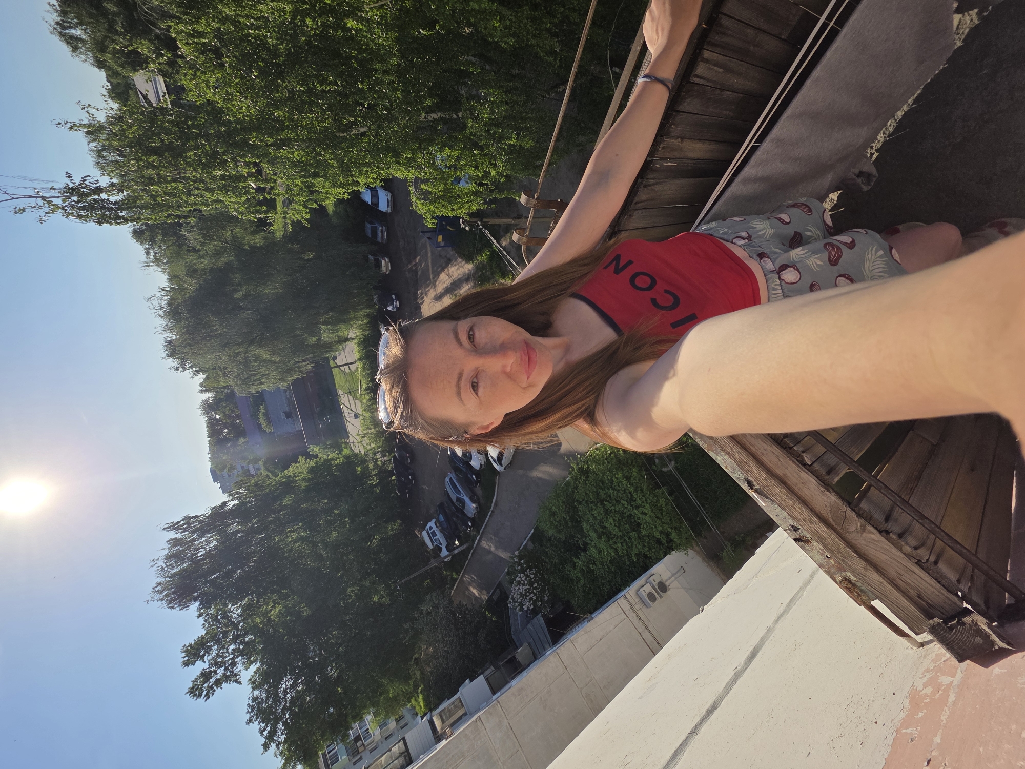 A selfie was taken in the city of Chaikovsky, Russia, depicting a fair-skinned woman with reddish-brown hair and sunglasses atop her head, wearing a red tank top and patterned shorts. She is on a roof or high structure with wooden edges and is holding the camera out at arm's length. Below, there's a tree-lined street with parked cars and buildings. The time of day seems to be sunny, as indicated by the bright sunlight and clear blue sky.