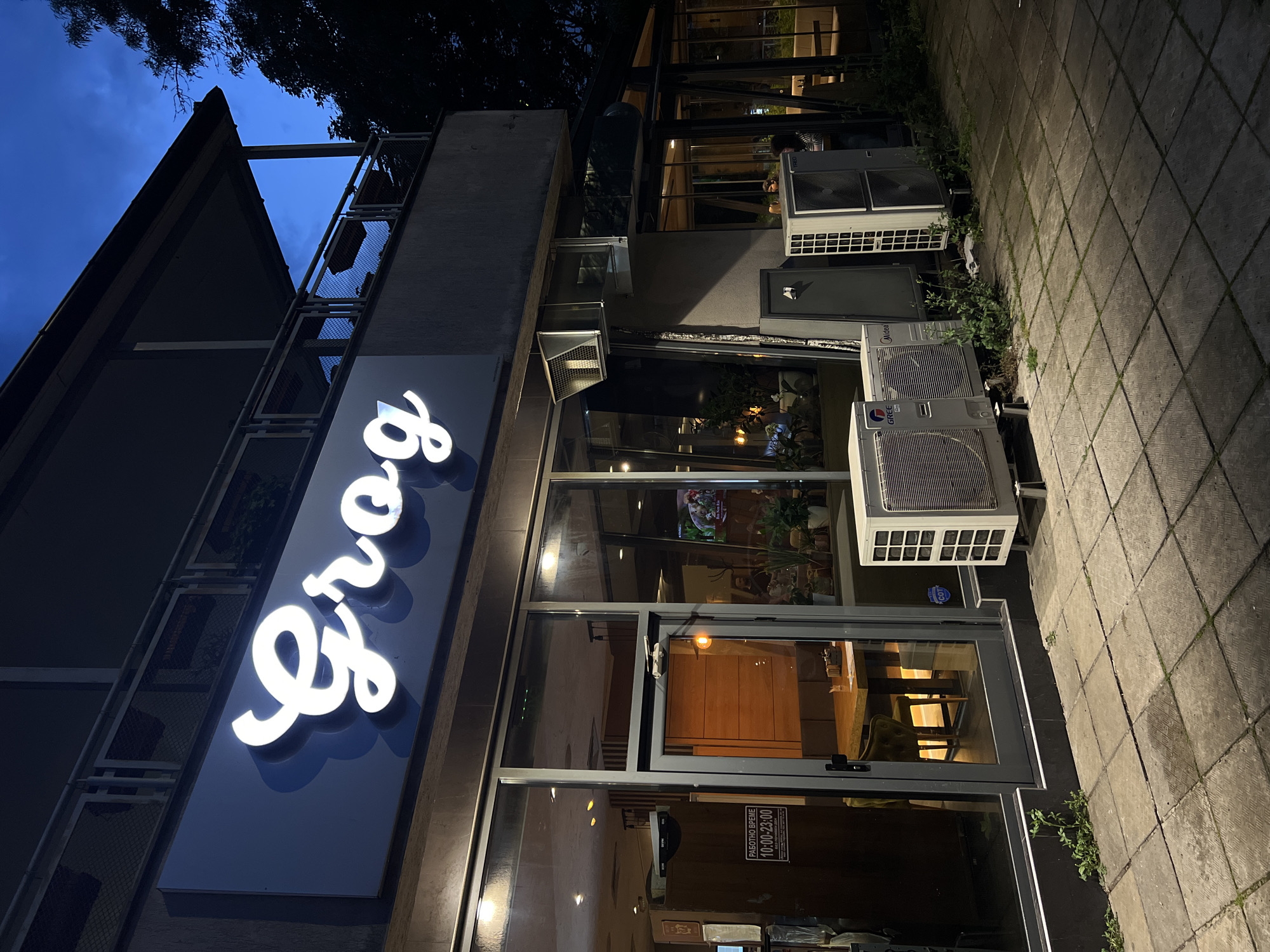 The image shows the exterior of a restaurant named "Greg". The building appears to be a modern design with large glass windows and a dark gray facade. The restaurant's sign, "Greg," is illuminated with white cursive letters. Air conditioning units are visible along the building's exterior. The setting is urban, with a paved sidewalk. A sign on the door shows Cyrillic text with the time 10:00 - 23:00, likely indicating the restaurant's operating hours. Based on this, and the sign's writing, this is likely the restaurant in Burgas, Bulgaria. The sky is dark, suggesting it is either evening or night.