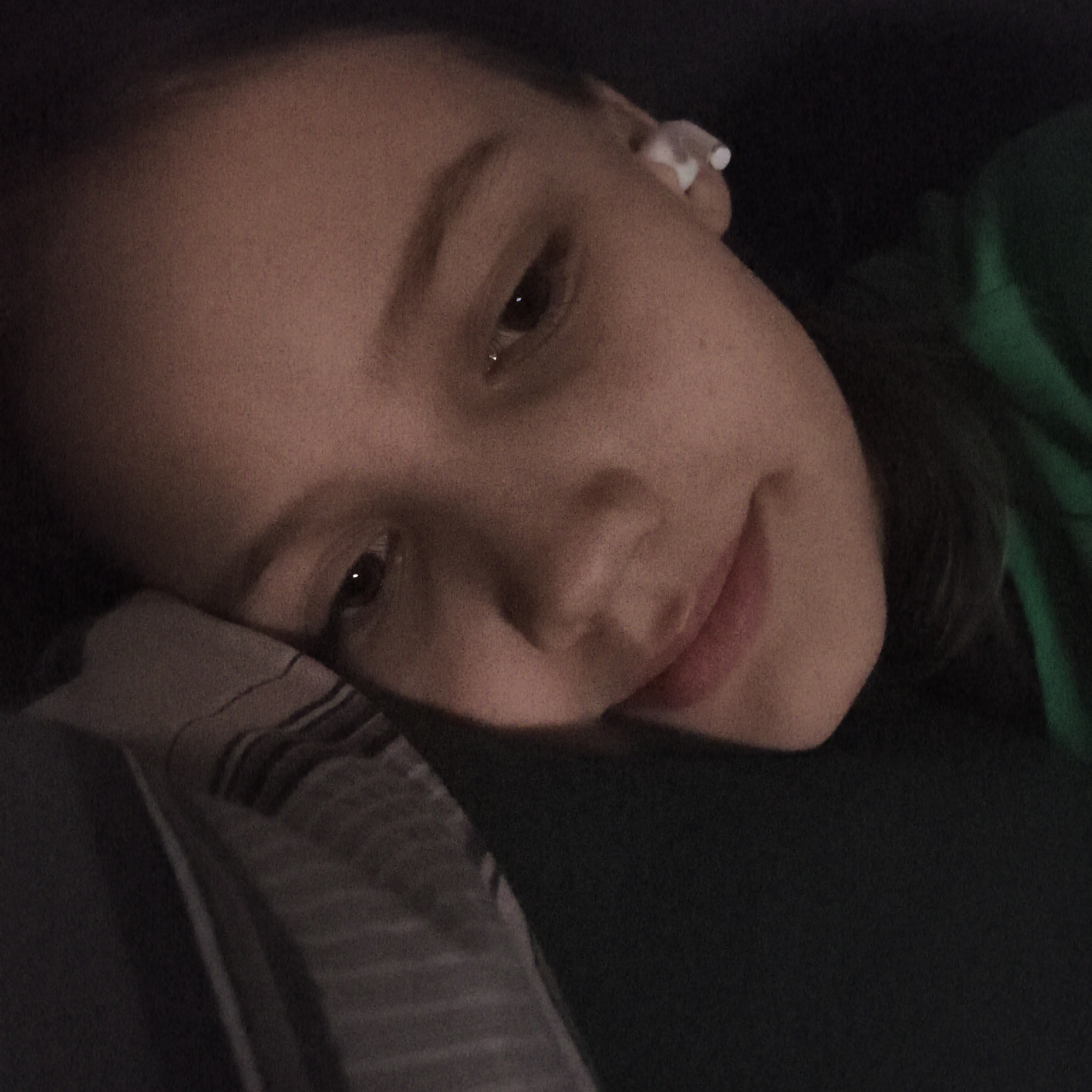 The image captures a close-up, dimly lit portrait of a young person lying down, likely in an indoor setting in Chaikovsky, Russia. The subject, appearing to be a teenager or young adult, rests their head on a patterned pillow or blanket, looking towards the camera with a soft, content expression. Their face is partially illuminated, creating a warm, intimate atmosphere, while much of the background remains dark.
A white earbud is visible in their right ear, suggesting they might be listening to music or engaged in a call. They are wearing what appears to be a dark green garment, possibly a hoodie or shirt, visible on the right side of the frame. The lighting conditions suggest it is either evening, night, or a dimly lit room, contributing to the serene mood. The image has a slightly grainy texture. No visible text is present. The overall scene conveys a moment of quiet relaxation or contemplation.
