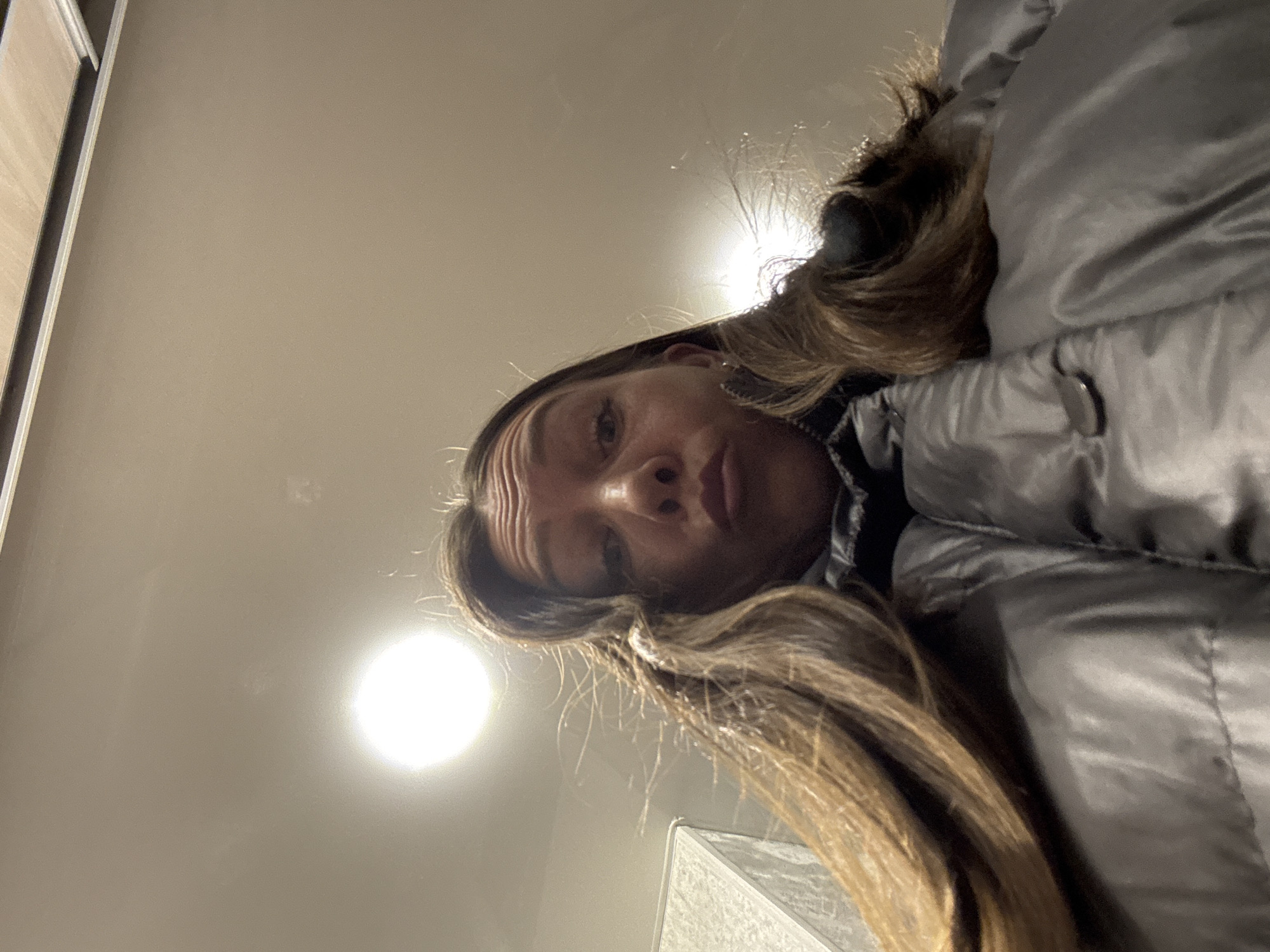 A low-angle shot shows a woman with long brown hair, wearing a grey puffy jacket. Above her is a light-colored ceiling illuminated by a bright light source. The location is in Chaikovsky, Russia.