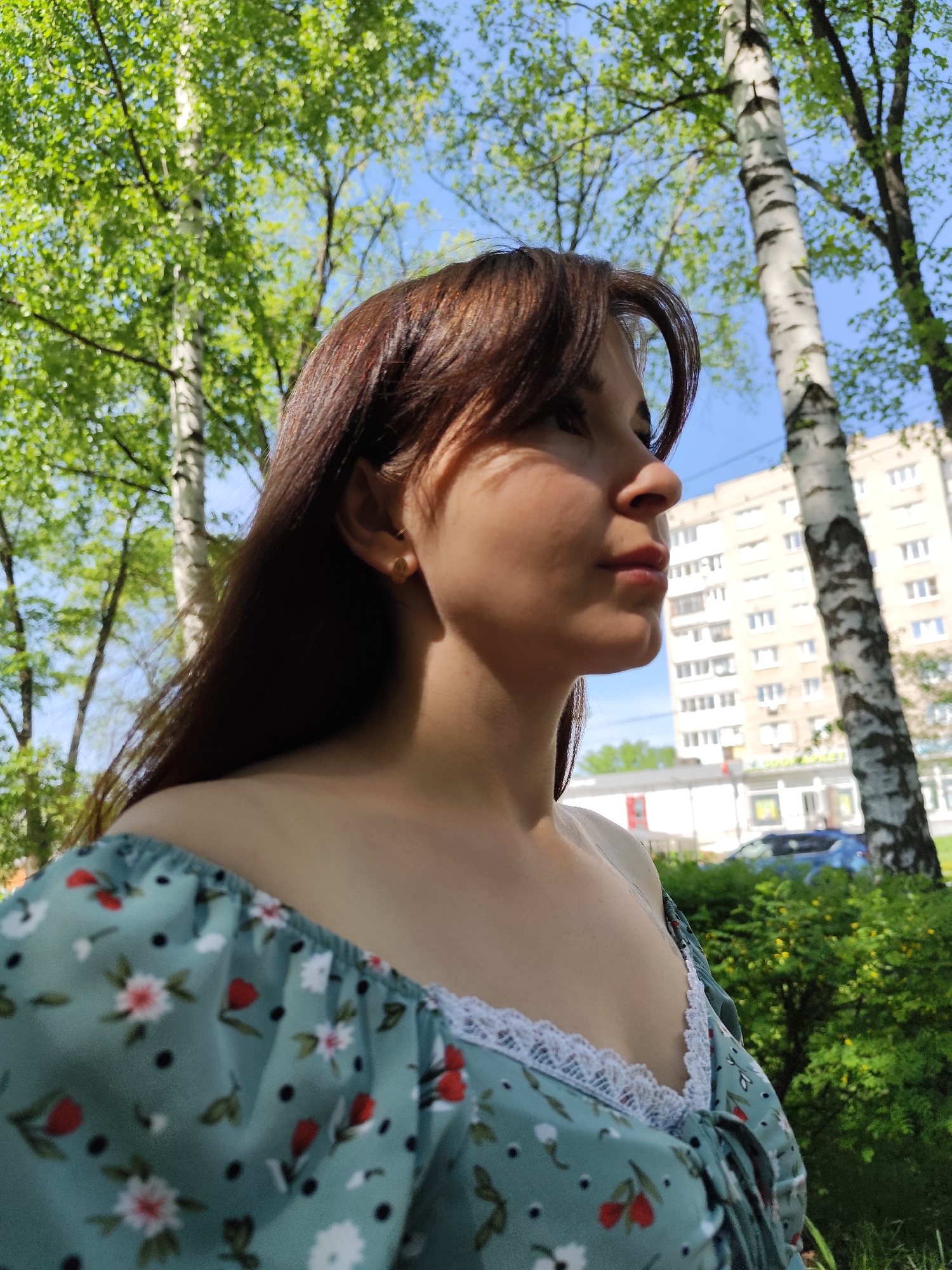 A young woman stands in a park in Chaikovsky, Russia, on a sunny day. The woman has long brown hair, is wearing a floral print dress with white lace trim, and has a gold earring. Her face is in profile, and the light casts shadows on her forehead and neck. Behind her, birch trees stand with their distinctive white bark and green leaves. In the background, a building with multiple windows can be seen, along with greenery and a glimpse of a blue car.