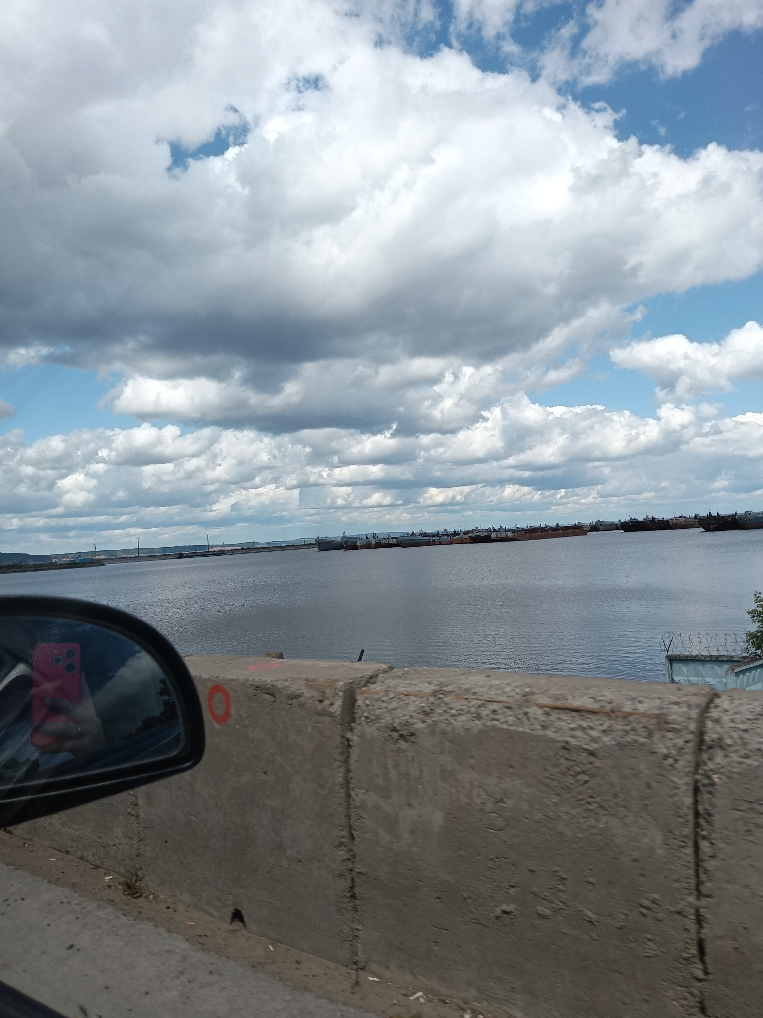 The image shows a view of the Kama River from a bridge in Chaikovsky, Russia. The foreground consists of a concrete bridge barrier with an orange circle painted on it. A car's side mirror is visible in the lower left corner, reflecting the photographer holding a pink cell phone. The Kama River fills the middle ground, with multiple ships or barges visible in the distance. The horizon is defined by a shoreline with industrial structures and vegetation. The sky is partly cloudy with a mix of dark gray and white clouds against a blue sky. Barbed wire is visible on a fence in the right middle ground near the river.