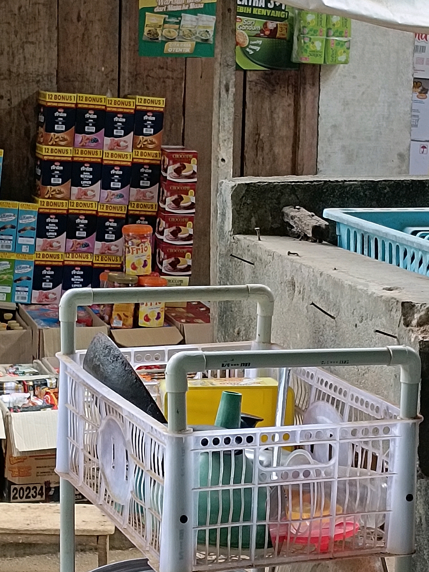 The image depicts a street market stall in Taman Cari, Indonesia. In the foreground, a white plastic shopping basket with a green handle is filled with various items, including a mortar and pestle, a yellow box, a green funnel, and other miscellaneous objects. Behind the basket, stacks of boxed snacks and cans are visible, some with "12 BONUS" labels. To the right, a blue plastic container sits on a concrete ledge. The background consists of wooden planks and a gray concrete wall. The scene appears to be outdoors, suggesting a commercial setting within the city. There are no people visible in the frame.