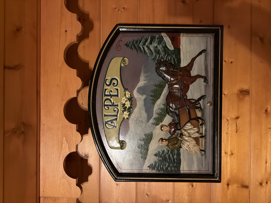 A wood-framed sign featuring a raised relief painting hangs on a wooden wall. The sign, labeled "ALPES," depicts a horse-drawn carriage carrying two passengers through a snowy mountain landscape with pine trees. The horse is rearing up, adding dynamism to the scene. The sign's frame is dark, contrasting with the lighter wood of the wall, which is adorned with decorative scalloped wood carvings on the left.