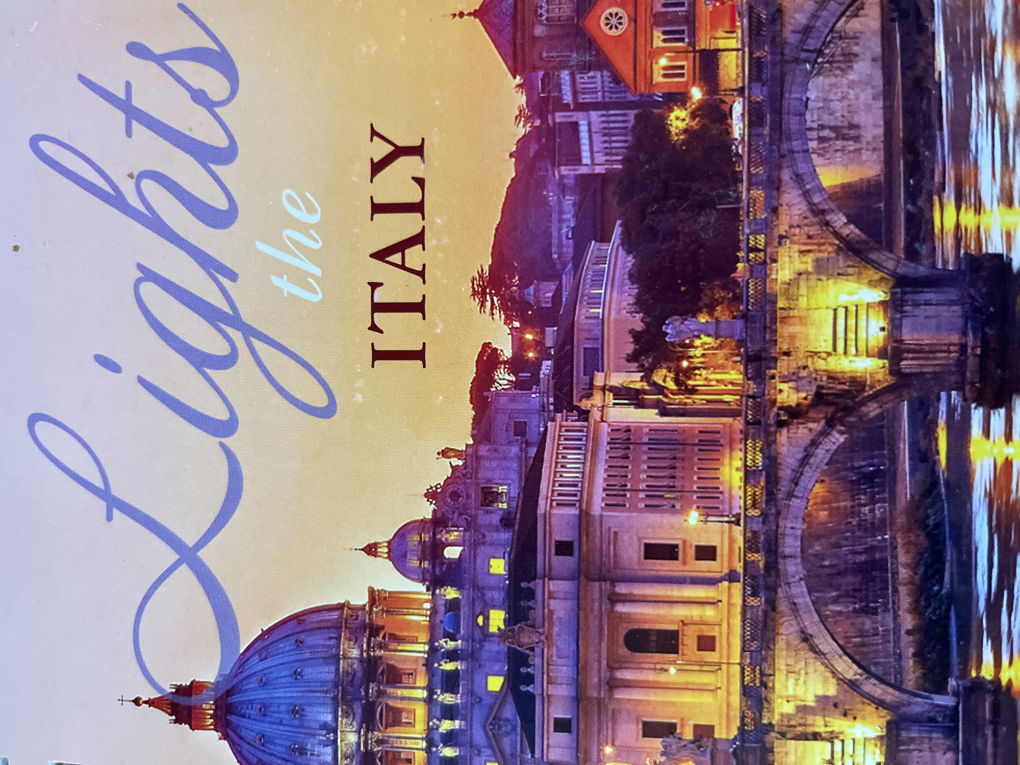 The image displays a vibrant nighttime or dusk cityscape, likely Rome, oriented vertically but capturing a horizontal landscape. A prominent large blue dome, characteristic of St. Peter's Basilica, is visible on the left side of the frame, surrounded by other classical-style buildings with illuminated windows.
A river, presumably the Tiber, flows across the middle and right sections of the image, reflecting the warm yellow and orange lights from the surrounding structures. Several arched stone bridges span the river, connecting the illuminated banks. The city's architecture ranges from grand, multi-story buildings with intricate details to a distinctive reddish-orange building with a circular window on the upper right. Artificial lights from streetlamps and building interiors cast a glowing ambiance over the scene. The sky is dark, emphasizing the brightness of the city lights.
Overlaid on the top left of the image is text. "Lights" is written in a large, flowing, light purple script. Below it, "the" appears in a smaller, light blue font. To the right, "ITALY" is displayed vertically in a bold, capitalized, dark brown serif font. No people or specific activities are discernible, with the focus remaining on the illuminated urban landscape and its reflections.