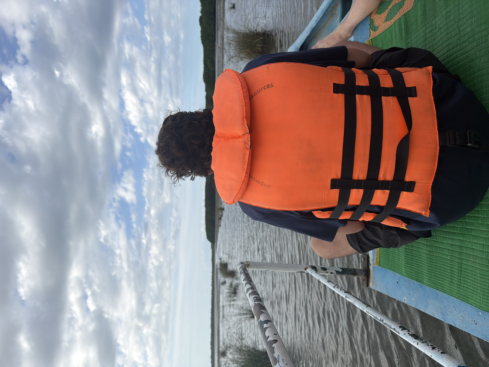 The image depicts a person from behind, wearing an orange life jacket over a dark blue shirt and black shorts, sitting on the edge of a blue and green boat. The boat is on a body of water with sparse reeds and vegetation. In the background, a treeline is visible under a cloudy sky. The scene appears to be a tranquil outing on a river or coastal area, possibly in Pottuvil, Sri Lanka, known for its diverse aquatic landscapes. The lighting suggests it is daytime, with diffused sunlight filtering through the clouds. The focus is on the individual and the immediate surroundings of the boat.
