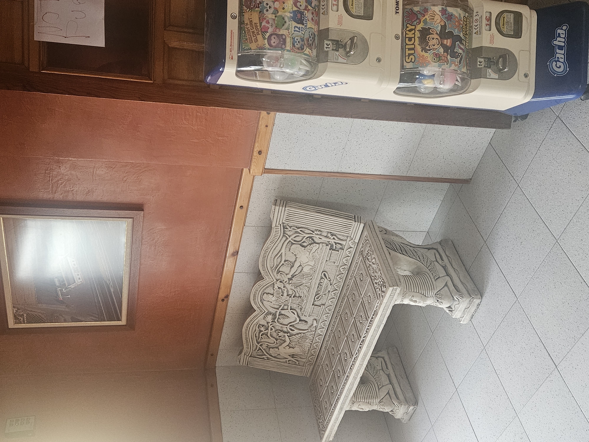The image depicts a section of a room with a combination of different elements. At the top is a vending machine filled with toy capsules, with "Gacha" prominently displayed. Underneath the machine, a richly decorated, ornate white stone or plaster table rests on a tiled floor. A framed picture hangs on the wall, with a brown frame against a wall painted in two tones of brown. The upper part of the wall is a darker, reddish-brown, while the lower part is a lighter, more muted brown. Above the vending machine, a piece of paper with the words "Pue" and "BBBB" written on it is taped to a wooden panel. The vending machine is on a wooden surface. The scene appears to be indoors in Albacete, Spain.