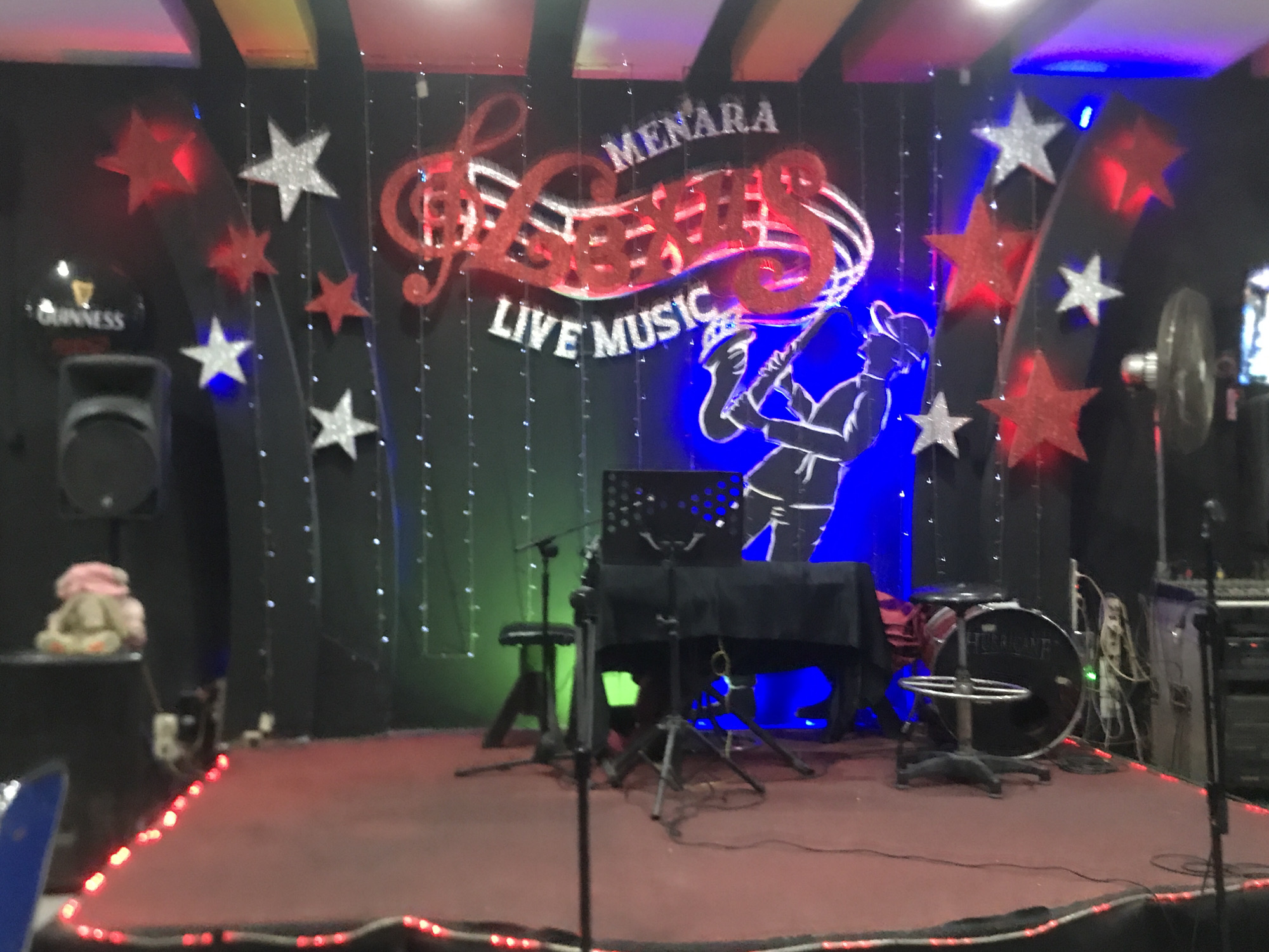 The image depicts a stage at Menara Lexus Live Music in Medan, Indonesia. The stage is set with a keyboard covered with a black cloth, drums, microphones, and a standing fan. Behind the stage is a black backdrop decorated with strings of lights, red and silver stars, and a lighted sign that reads "Menara Lexus Live Music." A speaker stands to the left, and another piece of equipment to the right. The stage itself is illuminated with red lights outlining its edge. A plush toy is visible on the speaker to the left.