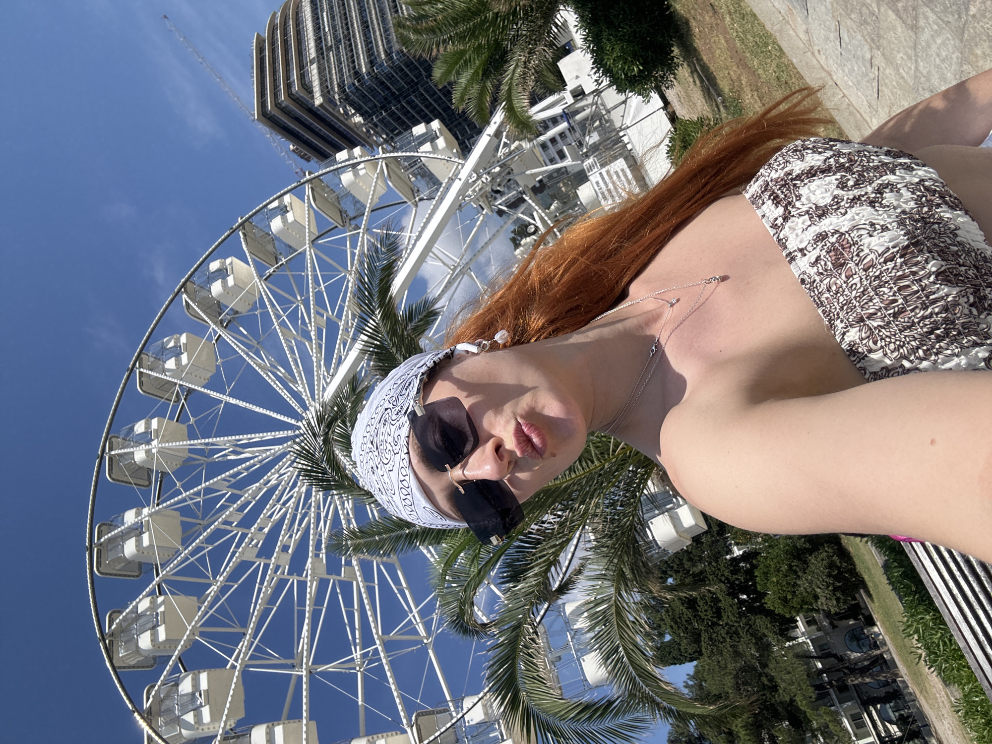 A selfie of a woman with red hair, wearing a white bandana and sunglasses, stands in front of a large Ferris wheel on a sunny day. The Ferris wheel is white with enclosed cars, set against a clear blue sky. In the background, there is a modern high-rise building and palm trees. The woman is wearing a top with a brown and white pattern and a silver necklace. The setting appears to be a recreational area in the city of Budva, Montenegro, based on the Ferris wheel and architecture visible.