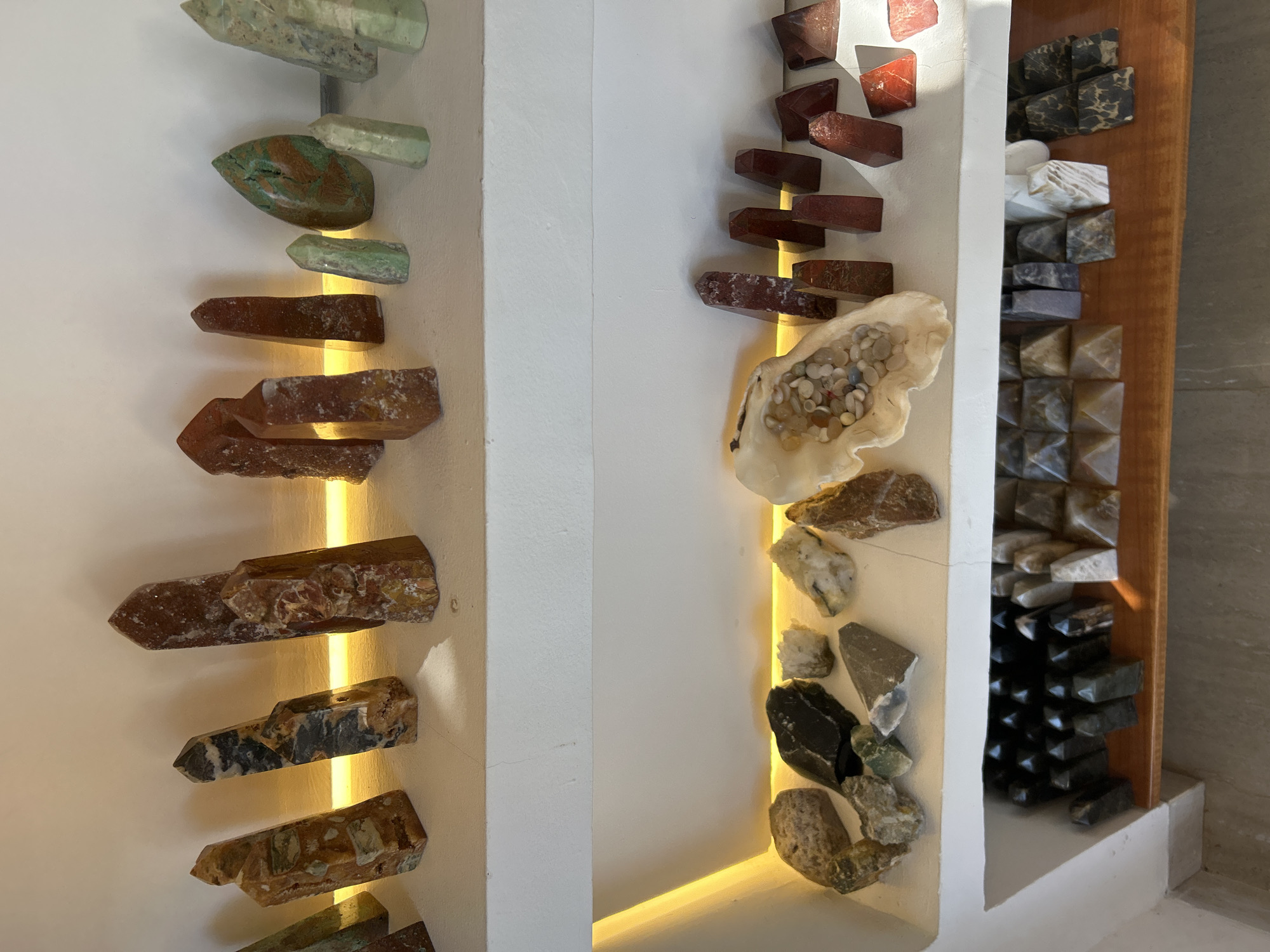 The image displays a collection of crystals and stones arranged on two white shelves. The top shelf features several tall, pointed crystals in various earthy tones, including reds, browns, and yellows, interspersed with a few lighter green and blue-green specimens. Some of these crystals have a rough, natural texture, while others appear polished.
The bottom shelf contains a variety of items. On the left side, there are rough, natural stones in shades of gray, black, white, and green. Towards the center, a large, irregular shell-like object is filled with small, colorful pebbles. To the right of the shell, a row of red, pointed crystals stands, followed by a collection of pyramid-shaped and other geometric stone objects in gray, white, and black, arranged in rows on a wooden shelf. The lighting appears to be artificial, with a warm glow illuminating the back of the bottom shelf. The background is a plain white wall.
This scene is located in a shop in Sukawati, Bali, Indonesia, known for its art and craft markets, which often feature items like these decorative stones and crystals.