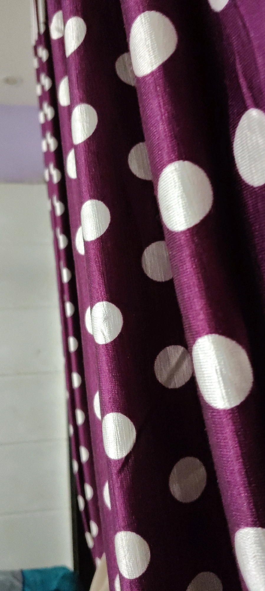 A close-up, vertical shot captures a portion of a purple curtain with a white polka dot pattern. The fabric is gathered and draped, creating folds and shadows that add depth to the image. The polka dots are circular and appear to be made of a lighter, possibly satin-like material that reflects light. The background is out of focus, showing a white wall and a glimpse of a light purple wall or ceiling. A dark vertical edge is visible on the left side of the frame, suggesting the curtain is hanging in a doorway or against a wall. The lighting appears to be natural, coming from the side, highlighting the texture of the curtain. The scene suggests an indoor setting, possibly a room in a residential or commercial space.