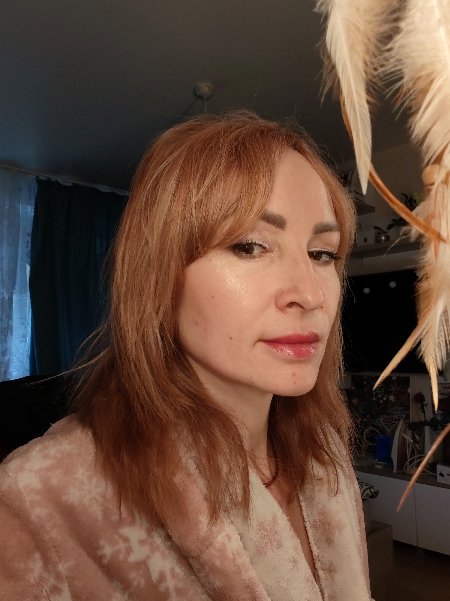 A woman with shoulder-length red hair and bangs is positioned in the foreground, wearing a pink and white patterned robe. She has light skin and is wearing makeup. Behind her, to the right, is a decorative object with light-colored feathers. Further in the background is a dark room with a curtain and some furniture and decor items. The photo was likely taken indoors and does not contain enough details to confirm that it was taken in Chaikovsky, Russia.