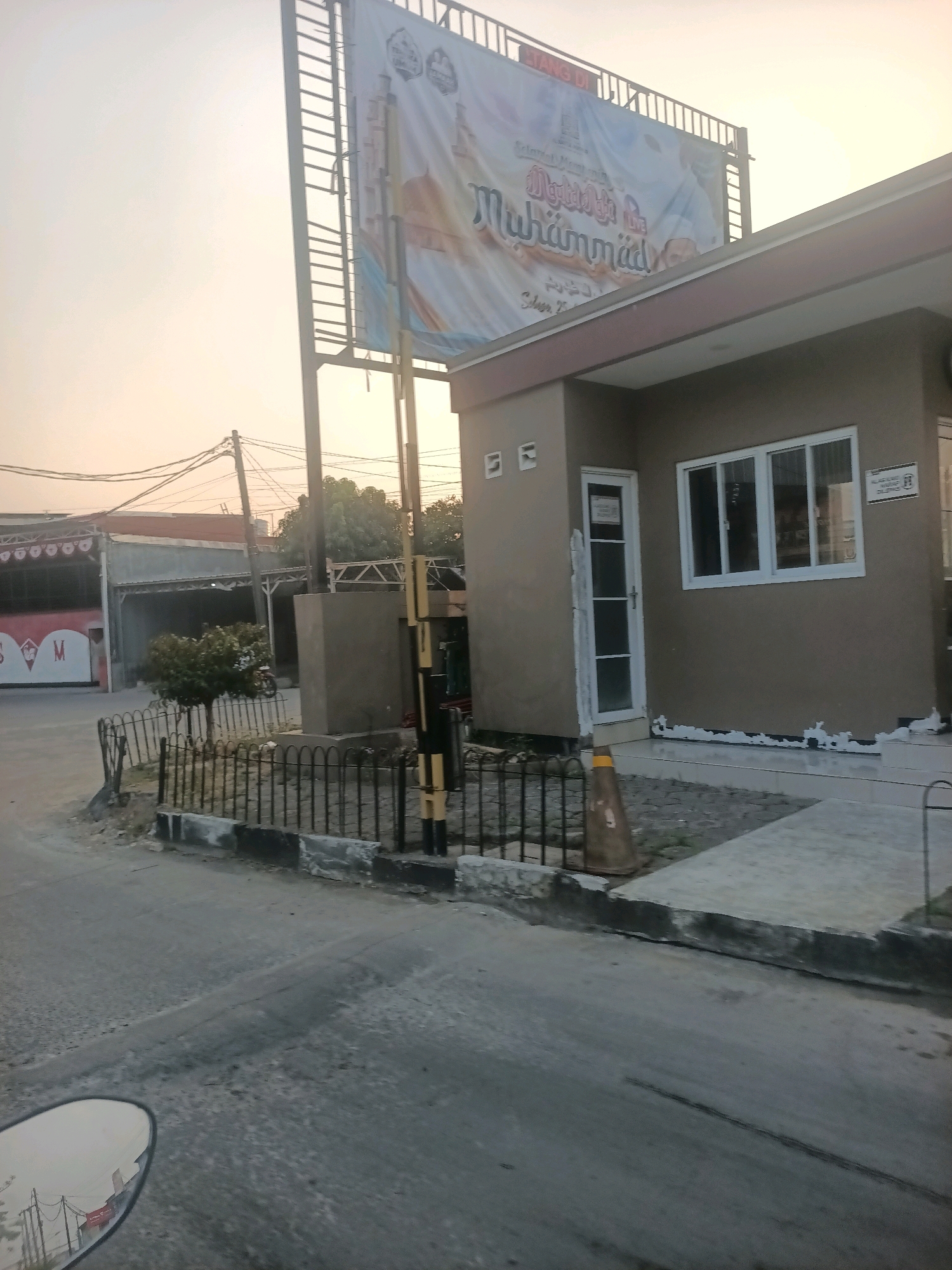 The image depicts a street scene in Wanajaya, Indonesia, at what appears to be dusk or dawn due to the soft, diffused light. In the foreground, a grey asphalt road is visible, with a motorcycle's side mirror reflecting a portion of the scene, including some buildings and power lines. A black metal fence runs along the side of the road, bordering a paved area. Behind the fence, a small, modern building with a tan facade stands, featuring a white-framed door and a large white-framed window with multiple panes. A traffic cone is placed on the paved area near the building. To the left of the building, a large billboard on a metal frame displays text and images, though the details are somewhat obscured by the lighting and resolution. Further in the background, power lines crisscross above a row of buildings, one of which has a red and white facade with circular logos and lettering. A small, bushy tree is situated in front of one of the background structures. A yellow and black striped pole, possibly part of a barrier system, is also visible near the building. The overall environment suggests an urban or semi-urban setting.