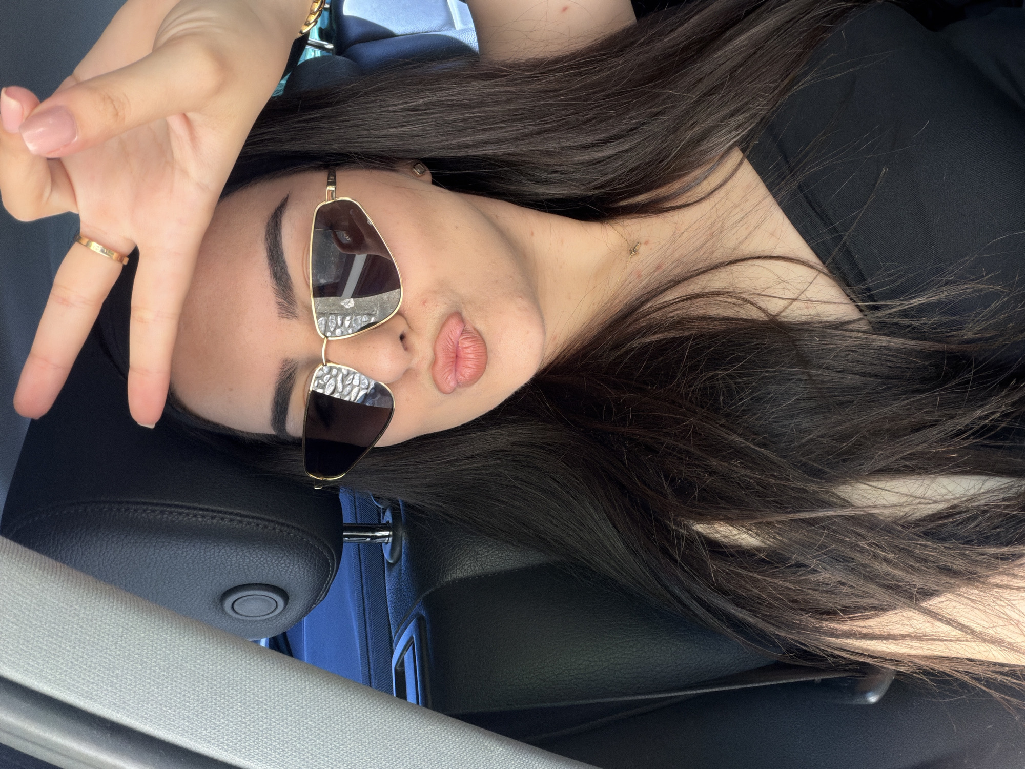 A young woman with long, dark hair is sitting in the passenger seat of a car, as indicated by the black leather headrest and a grey fabric-covered pillar. She is wearing a black strapless top, sunglasses with gold frames, and a thin gold ring on her right hand. She is making a peace sign with her right hand, which is positioned near her forehead. Her lips are pursed, and she appears to be looking at the camera. The background is blurred, but suggests an outdoor setting, possibly a street or a parked area, given the reflection in her sunglasses which appears to be buildings and trees. There is a glimpse of a thin gold necklace with a charm around her neck. The lighting suggests it is daytime.