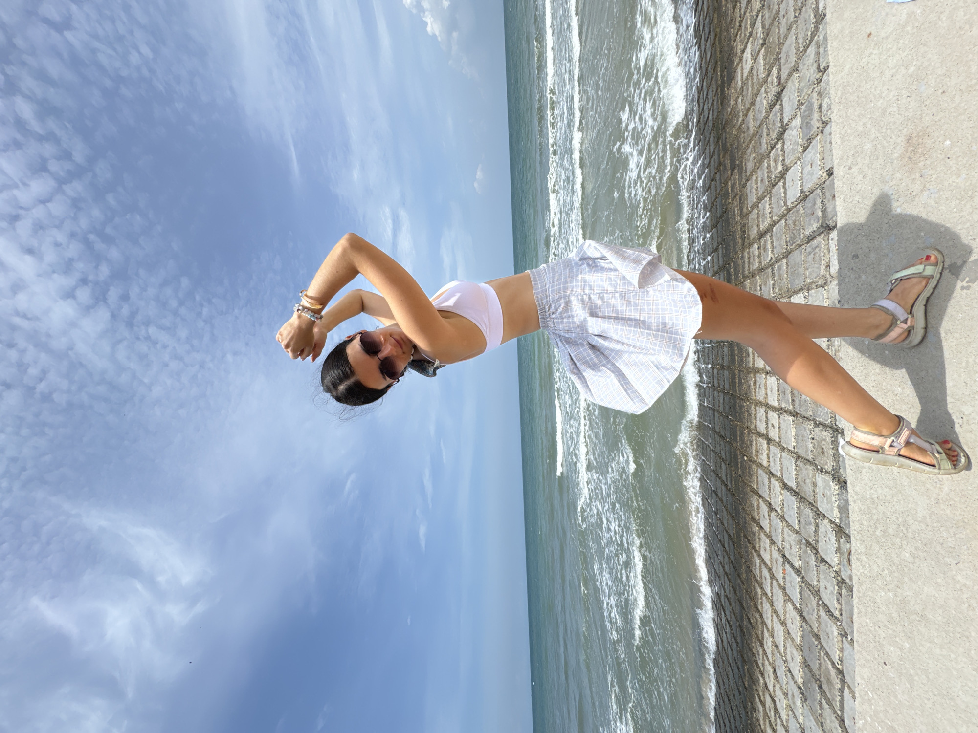 A young woman stands on a concrete walkway by the sea. She is wearing a white bikini top, a plaid skirt, sunglasses, and sandals. The sea is choppy with waves and a blue sky with scattered clouds is above. The walkway is made of textured concrete blocks. The location is Phu Thuy Ward, Vietnam.
