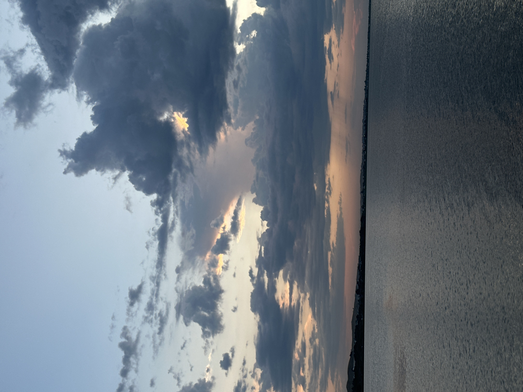 The image depicts a serene twilight scene over a body of water, likely a lake or bay. The sky is a canvas of soft blues and grays, transitioning into hues of orange and pink near the horizon, indicating the sun has recently set. Dramatic, dark clouds dominate the upper portion of the sky, with pockets of warm light breaking through in places. Below the sky, a dark, silhouetted shoreline stretches across the frame, dotted with what appear to be low-rise buildings. The water in the foreground is calm, with subtle ripples reflecting the muted colors of the sky. There are no people, animals, or specific landmarks visible that would definitively pinpoint the location within Kokshetau, Kazakhstan, but the overall environment suggests a lakeside or coastal urban fringe.