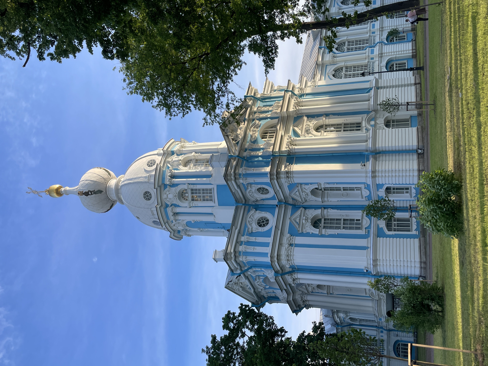 The image displays the Smolny Cathedral in Saint Petersburg, Russia, under a clear blue sky. The cathedral, constructed in a Baroque architectural style, features a white and light blue facade with numerous columns, ornate window frames, and decorative elements. A large white central dome with circular windows is topped by a smaller white dome and a golden spire culminating in a cross. The building's visible side is illuminated by direct sunlight, casting distinct shadows.
In the foreground, a neatly maintained green grassy lawn is present, interspersed with small bushes and several young trees supported by stakes. Mature green trees flank both sides of the cathedral, their foliage extending into the upper portions of the frame. The sky is bright blue with minimal, faint white cloud wisps visible in the upper left quadrant, and a faint disk, possibly the moon, is discernible in the upper center against the blue sky.
On the far right, near the base of the cathedral and a mature tree, two individuals are seen walking away from the viewer on a paved path. One person wears lighter-colored clothing, and the other wears darker attire. A dark lamppost stands on the lawn near them.