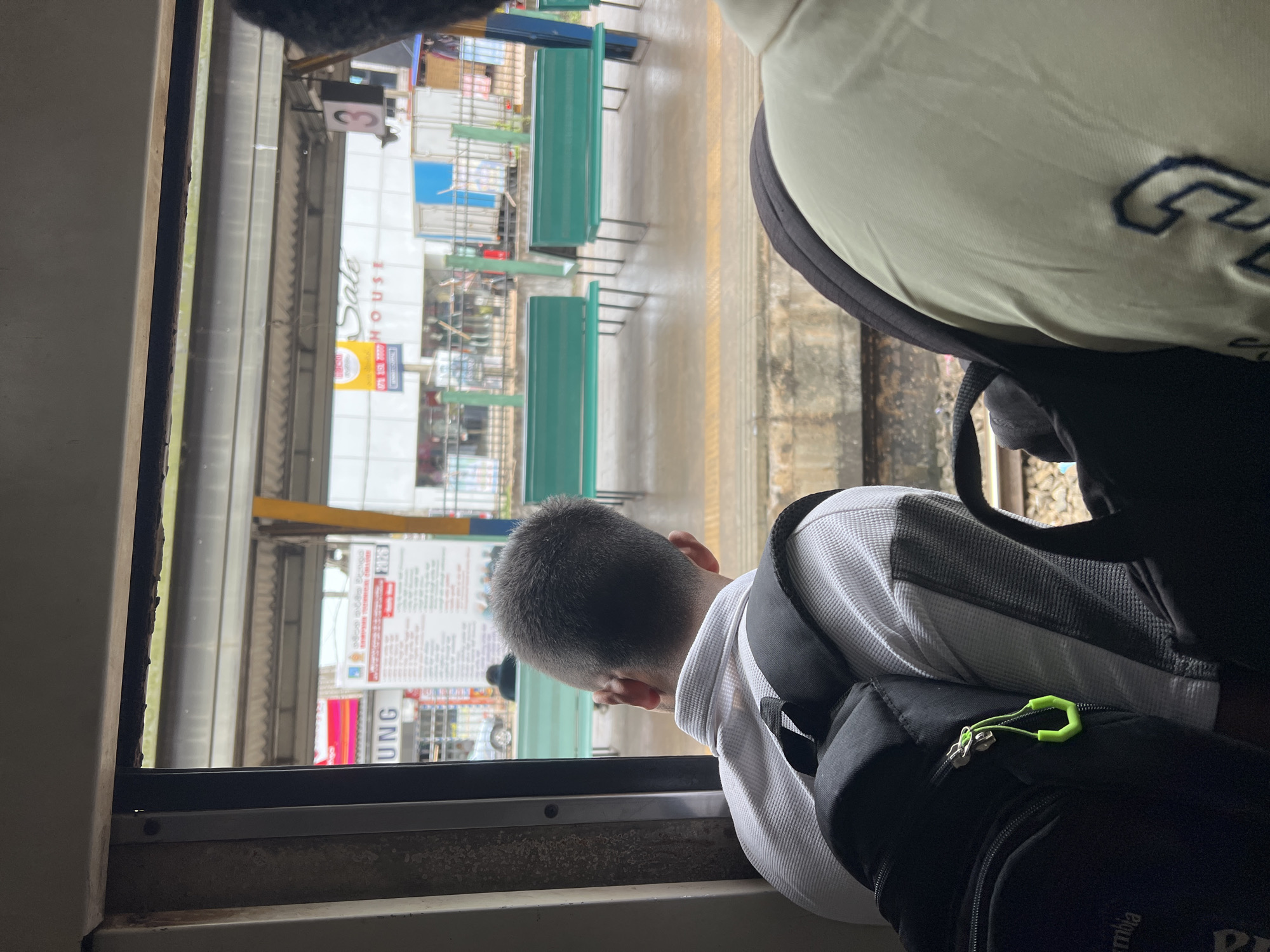 The image depicts a view from inside a train carriage, looking out a window at a train station platform. Two individuals are seated in the carriage, their backs to the camera. The person in the foreground on the left is wearing a white collared shirt and has a black backpack on. The person on the right is wearing a light-colored shirt with a navy blue logo. Outside the window, the platform is visible with green benches, a covered walkway, and buildings in the background. A sign on one of the buildings reads "SALE HOUSE". There are also several people on the platform. The scene appears to be in the city of Gampaha, Sri Lanka, suggested by the context.