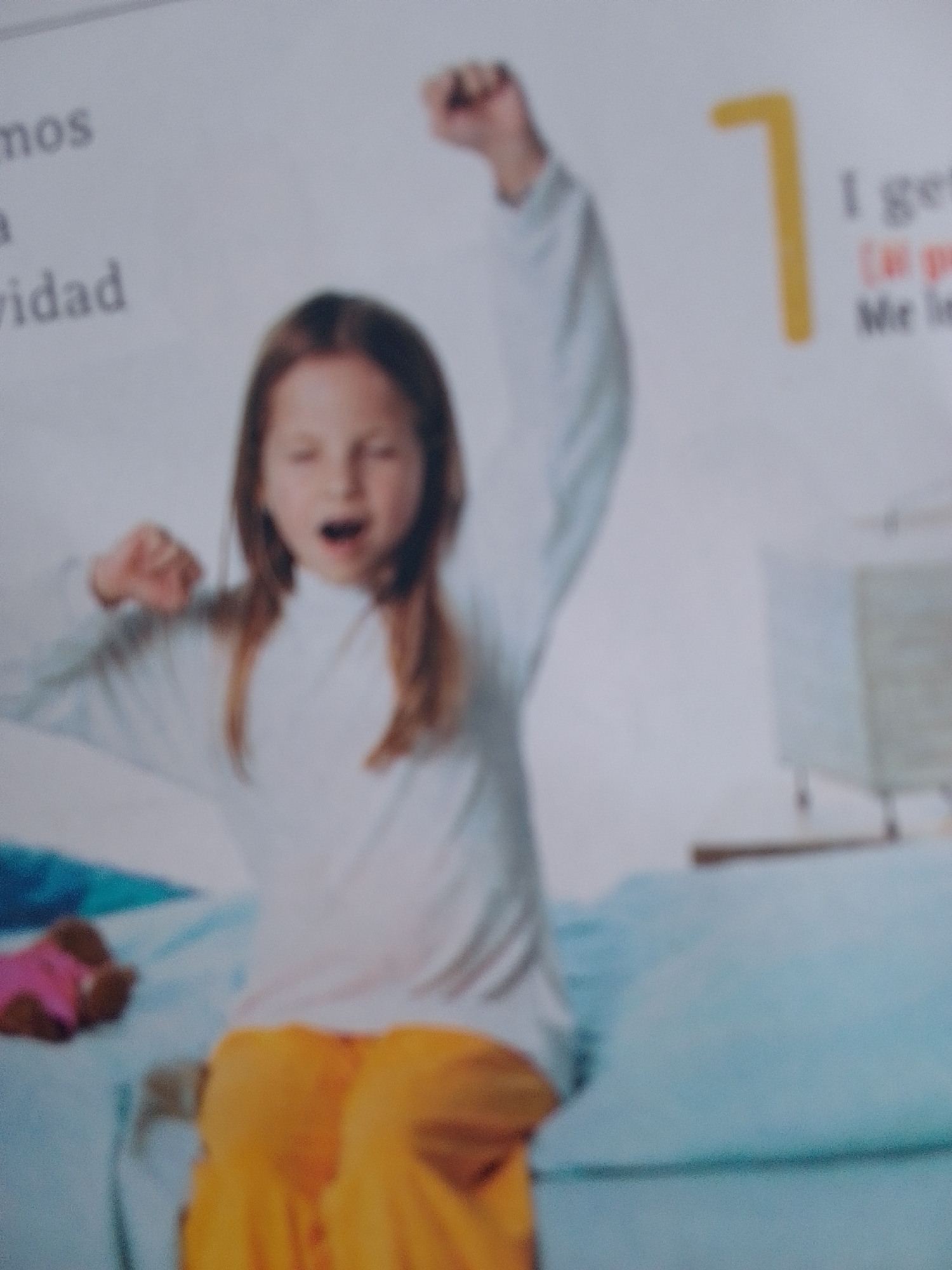This media file features a young girl with brown hair, appearing to be around 6-8 years old, yawning or stretching. She is wearing a white long-sleeved shirt and bright orange pants. The girl is positioned on what appears to be a bed, with light blue bedding. To the left of the girl, a small stuffed animal with pink and brown colors is visible on the bed. In the background, a lamp with a light-colored shade is present.
On the left side of the image, there is visible text that reads "mos", "a", and "vidad". On the right side, there is a large yellow number "7" and some text that appears to be "I get" followed by other words that are difficult to discern due to the image quality. The lighting suggests it could be daytime. The overall scene conveys a sense of waking up or stretching, potentially in a bedroom setting in La Tagua, Venezuela. The girl's expression is one of a yawn or stretch.