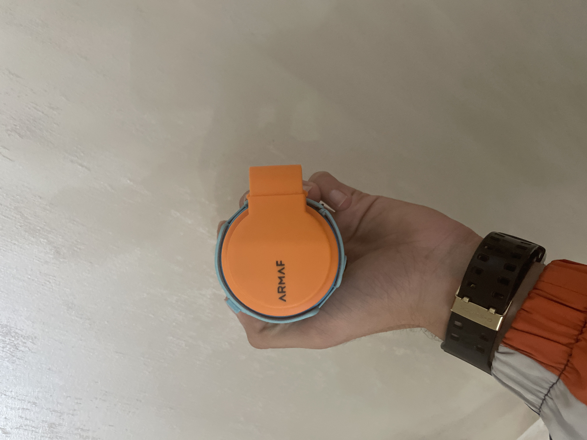 A human right hand is centrally positioned, holding a compact, cylindrical object. The object features a matte orange circular top with the word 'ARMAAF' printed in black capital letters. A small, flat orange tab or handle extends from the right side of the orange top. The perimeter of the object is light blue. Below the hand, a wrist is visible, adorned with a dark, possibly black, wide-strapped watch featuring rectangular perforations and a gold-colored clasp. The cuff of a jacket is visible on the lower left, exhibiting segments of orange, white, and a darker color, likely blue or grey. The background consists of a uniformly lit, textured wall in a light, neutral tone, possibly off-white or beige. This scene is located in the city of Ihtiman, Bulgaria.