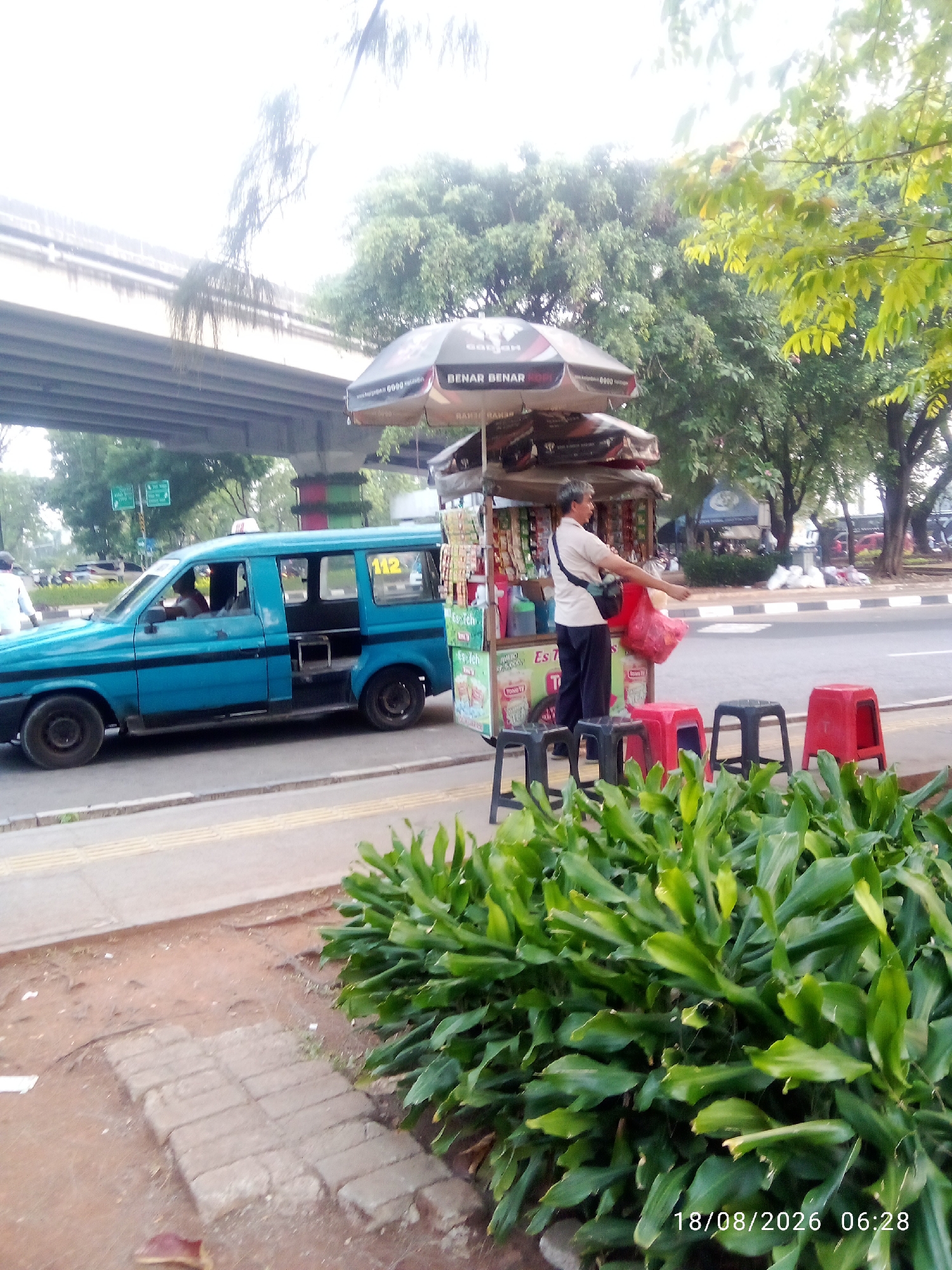 In a street scene in East Jakarta, Indonesia, a blue taxi with the number "112" is parked. In front of a food cart, a man in a light-colored shirt and dark pants is holding a red plastic bag, likely a customer. The food cart is covered by an umbrella with the words "BENAR BENAR" visible. Several small stools, some red and some black, are arranged in front of the cart. Lush green foliage is in the foreground. An elevated road or bridge is visible in the background, along with trees and other vehicles. The date and time stamp "18/08/2026 06:28" is present in the lower right corner.