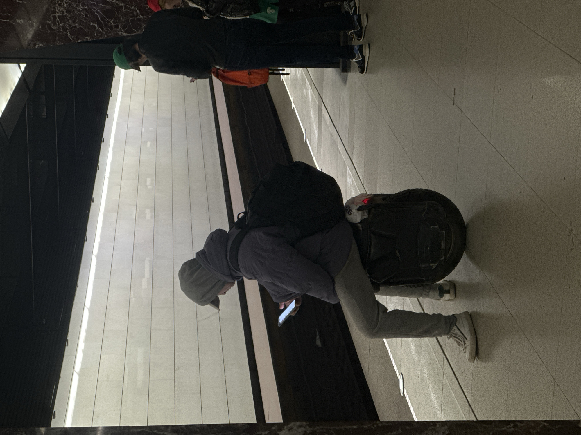 A man wearing a grey hoodie and grey sweatpants is standing with an electric unicycle, looking at a smartphone. He is carrying a black backpack. In the background, two other people are standing, one wearing a green hat and another with a red hat. The environment appears to be a modern interior space with tiled walls and flooring, potentially a metro station. Based on the architectural style, it could be located in Moscow, Russia.