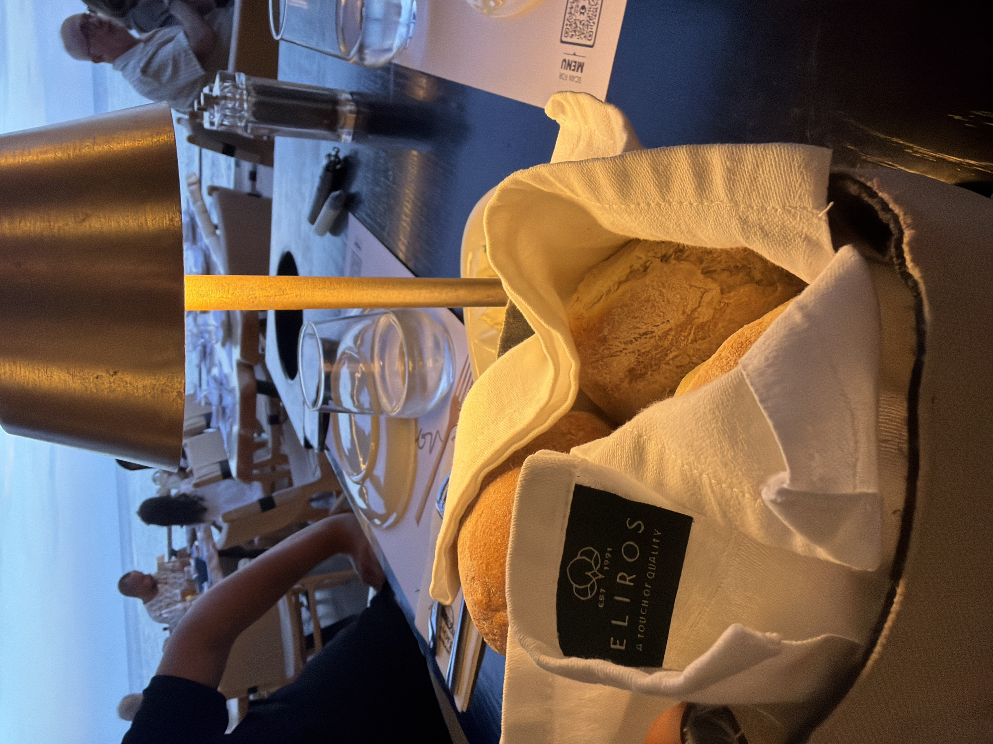 The image captures a relaxed evening dining scene in Makri, Greece, presented sideways (rotated 90 degrees clockwise). In the foreground, a wicker bread basket lined with a white fabric napkin holds several golden-brown bread rolls. A black label sewn onto the napkin displays the brand "ELIROS," with "EST 1991" and "A TOUCH OF QUALITY" beneath it.
The table, dark in color, is set with clear drinking glasses and a minimalist salt and pepper shaker set. A small, golden-hued table lamp provides a warm, localized glow. A white card featuring an upside-down QR code and the text "SCAN FOR MENU" and "MENU" is also visible. Part of a person's arm and hand are seen on the left side of the table.
The background reveals other diners seated at tables, suggesting a restaurant or resort environment. Beyond the dining area, a light blue sky extends to the horizon, indicative of an outdoor setting, likely at dusk or early evening. The ambient lighting and the visible sky imply an open-air or beachfront restaurant. The overall impression is one of a calm and pleasant meal.