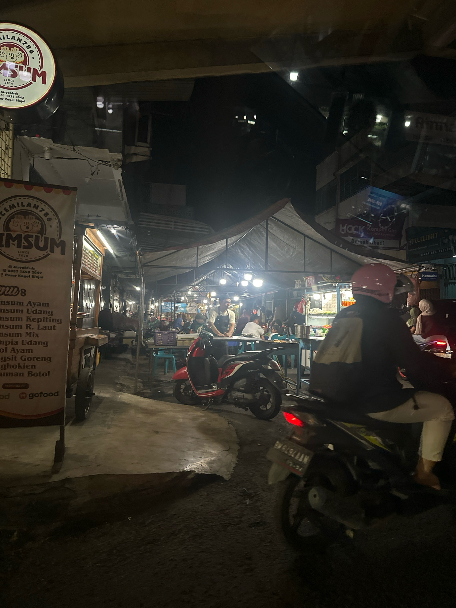 The image depicts a nighttime scene at a street food market, likely located in the City of Binjai, Indonesia. In the foreground, a motorcycle with a rider wearing a pink helmet is visible, partially obscuring the view of the market stalls. The market is set up under temporary awnings, illuminated by various lights. Several food stalls are visible, with people seated at tables and standing around. One stall prominently features a sign for "CEMILAN786 DIMSUM", indicating the sale of dim sum. The ground appears to be paved, with some unevenness. Reflections on the surface suggest the presence of water or a wet surface. The overall atmosphere is that of a bustling, casual dining environment at night.