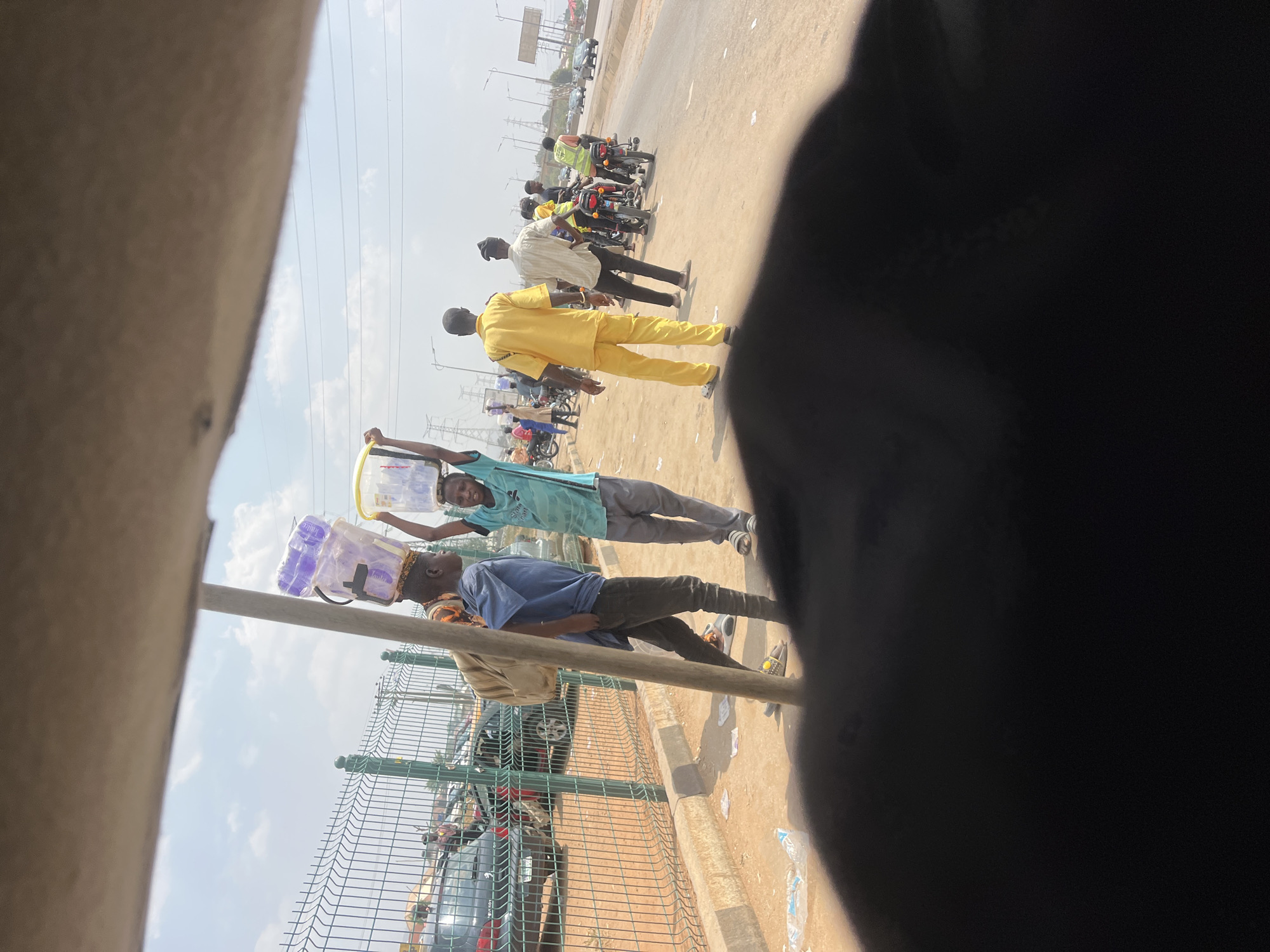 The scene is a roadside in Galadimawa Village, Nigeria. Two men are carrying goods on their heads, walking along the roadside next to a fence. One man carries a container filled with small purple bags, while the other carries a clear bucket. Further down the road, a group of men are walking in the same direction along the roadside, some wearing distinctive yellow or white shirts. In the background, there is a row of parked motorcycles on the shoulder of the road. Cars are visible on the road. The sky is overcast. The image seems to be taken from inside a vehicle, as a part of the interior is visible in the foreground.