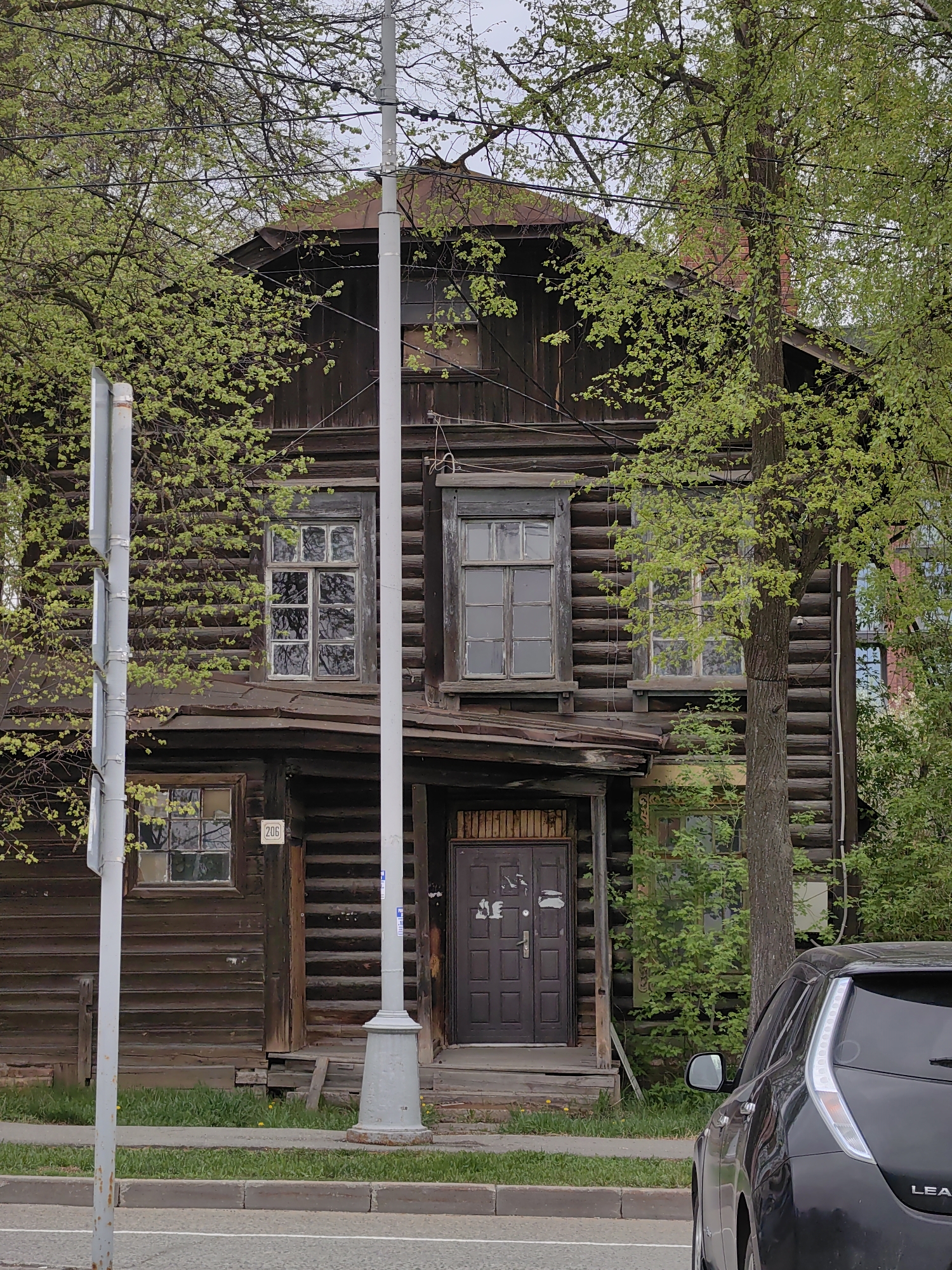 The image shows a two-story wooden house in Izhevsk, Russia. The house is constructed from dark brown logs and has a metal roof. The address "206" is visible on the front of the house. There are several windows with multiple panes, some of which appear to be boarded up or in disrepair. A grey utility pole stands in front of the house, partially obscuring the view. The house is surrounded by trees with green leaves, indicating it is likely spring or summer. A black car is parked on the right side of the frame, partially blocking the view of the house. The scene appears to be taken from across the street. The overall impression is of an old, possibly abandoned, house in a city setting.
