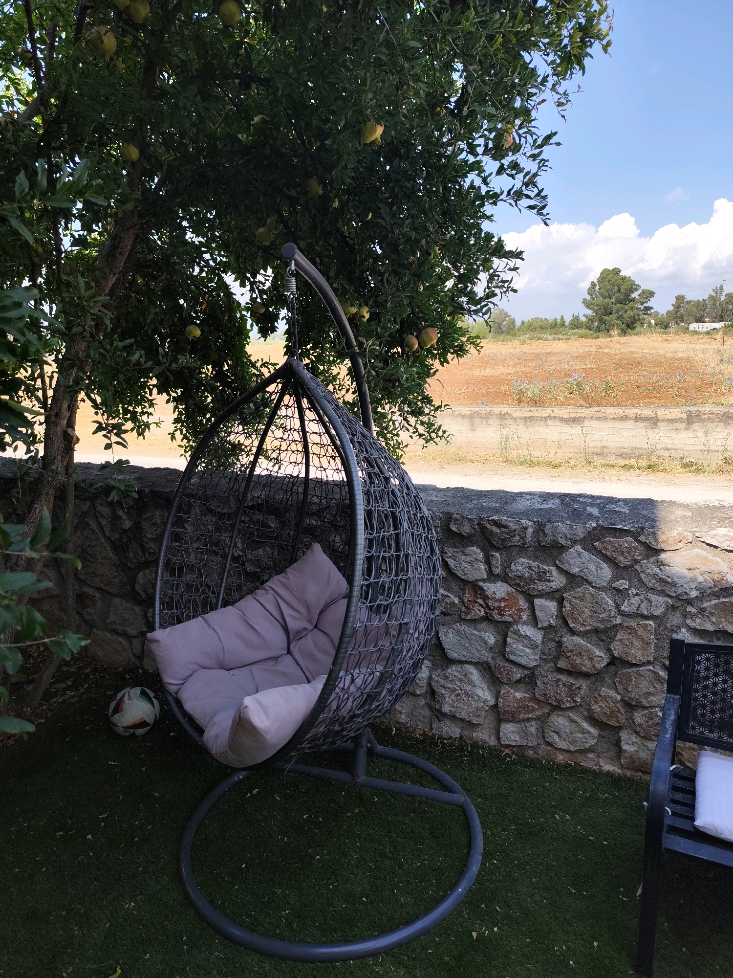 The image depicts an outdoor scene with a hanging egg chair on a circular base, situated on artificial grass. The chair contains a light pink cushion. To the left of the chair, there is a soccer ball. Behind the chair, a stone wall runs horizontally across the frame. Above the wall, a tree with green leaves and some yellow fruits is visible. The background shows a dry, reddish-brown field with sparse vegetation and some distant trees and buildings under a bright blue sky with scattered white clouds. The overall impression is one of a garden or patio area. The location is the Municipal Unit of Nafplio, Greece.
