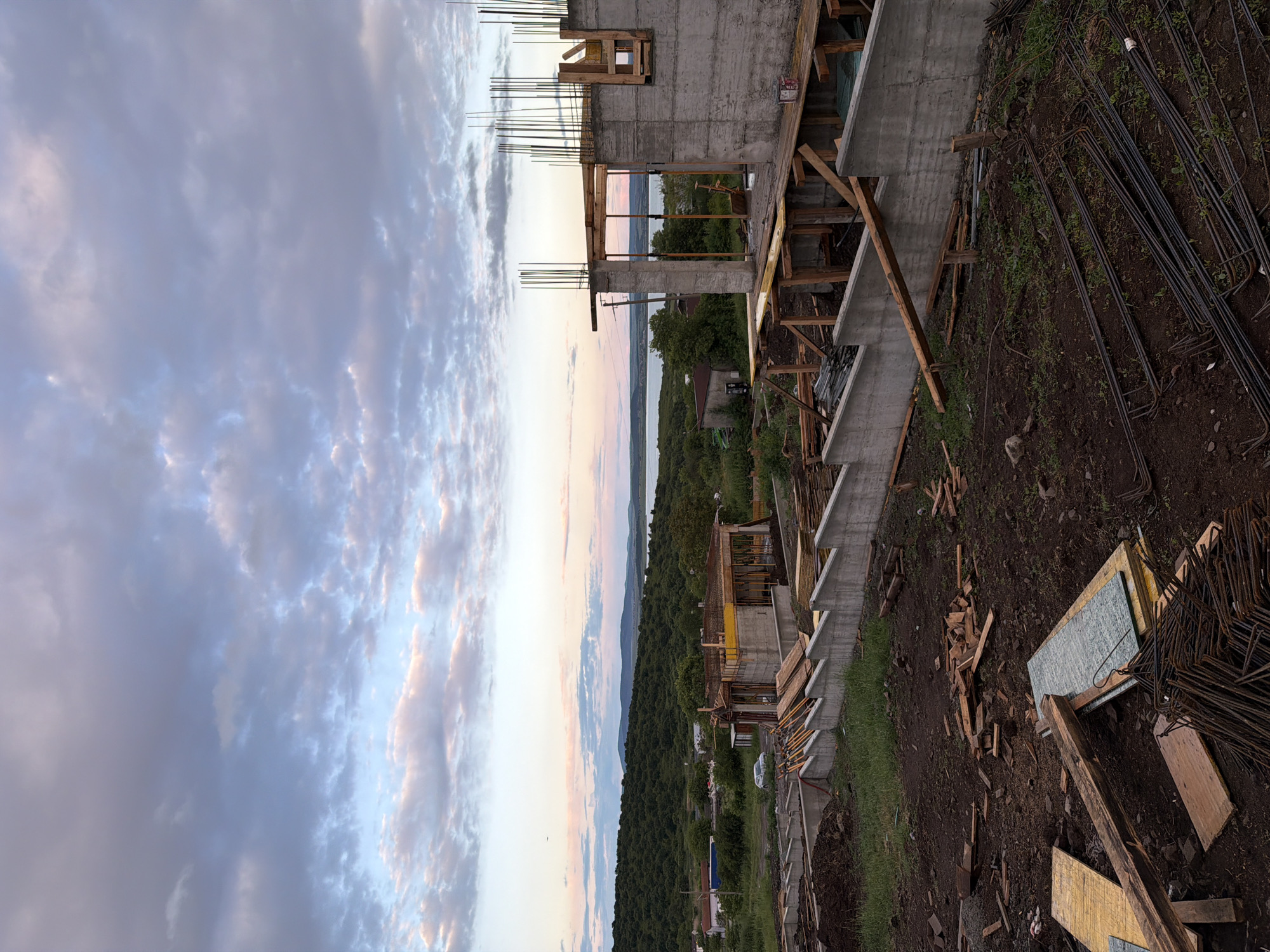 The image shows a construction site in Cherni Vrah, Bulgaria, against a backdrop of a sunset-lit sky. The partially constructed building features concrete walls with exposed rebar and wooden framework for windows and doors. The surrounding landscape includes a forested area leading to a body of water in the distance. Construction materials like lumber and rebar are scattered on the ground around the building's base. There are no people visible in the scene.