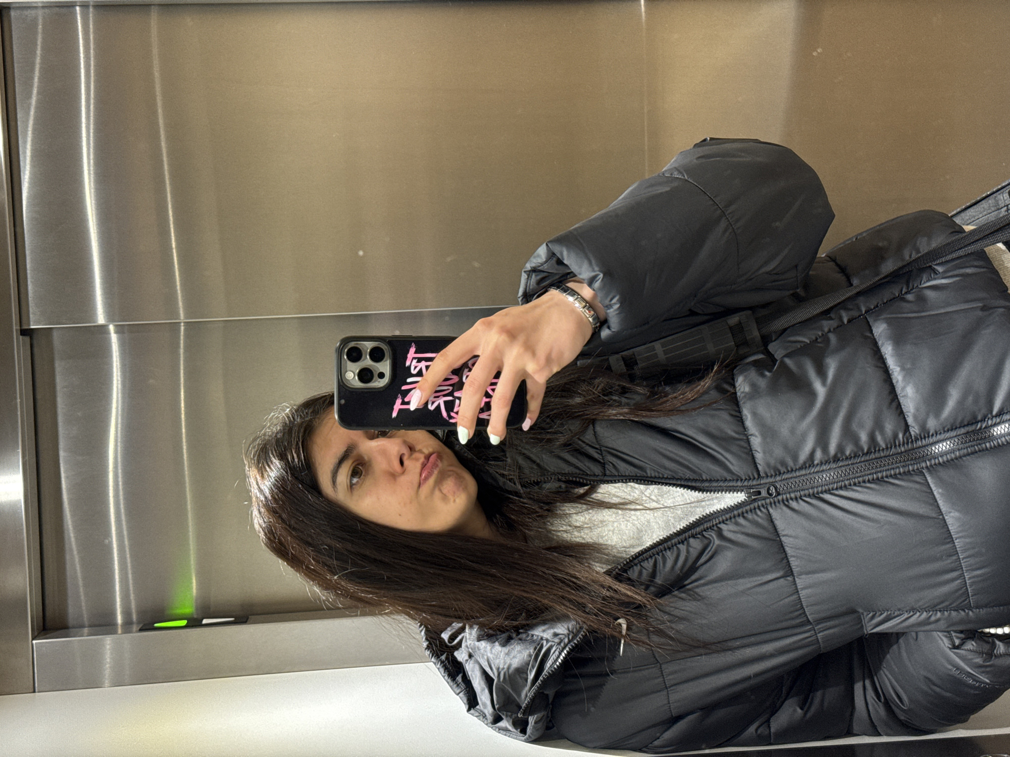 A woman stands in front of a silver-colored elevator, holding her phone in her right hand to take a selfie. She has long, dark brown hair and is wearing a black puffy jacket over a white sweater. Her phone case is black with pink writing. The elevator has a green light indicator visible on its control panel. The location is likely inside a building in Sofia, Bulgaria, though that is not definitively indicated in the image.