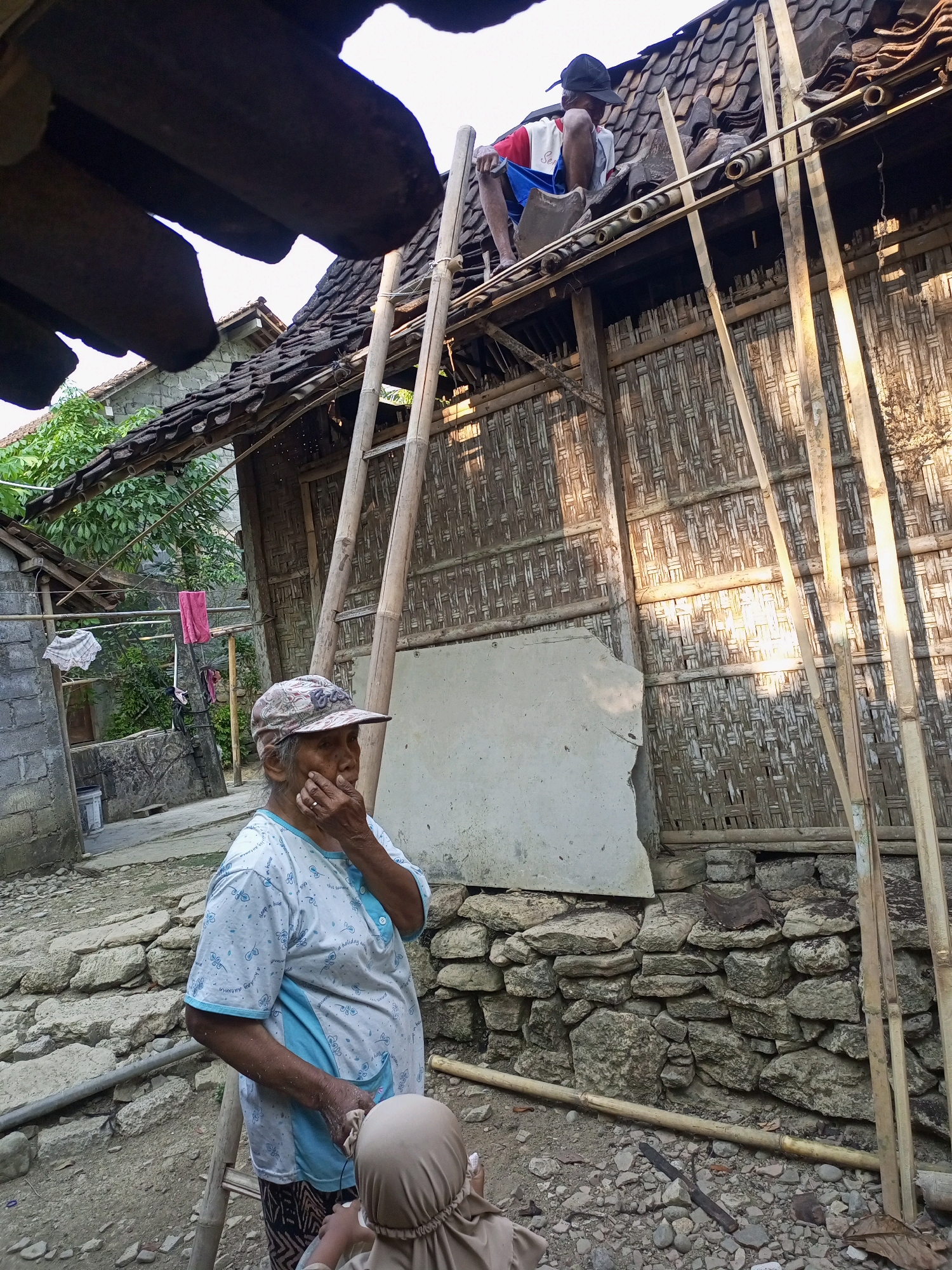 In Nanggulan, Indonesia, an elderly woman wearing a patterned cap and a blue and white shirt stands in the foreground, her hand touching her chin as she looks to her left. She is near a bamboo ladder leaning against a building. In the background, a younger person wearing a black cap, red and white shirt, and blue shorts sits on the roof of a building, working with what appears to be roof tiles. The building has walls made of woven bamboo and a tiled roof, and is partially covered by a makeshift scaffolding of bamboo poles. The scene is set in a rural or semi-rural environment, with visible stone walls, dirt ground, and foliage.