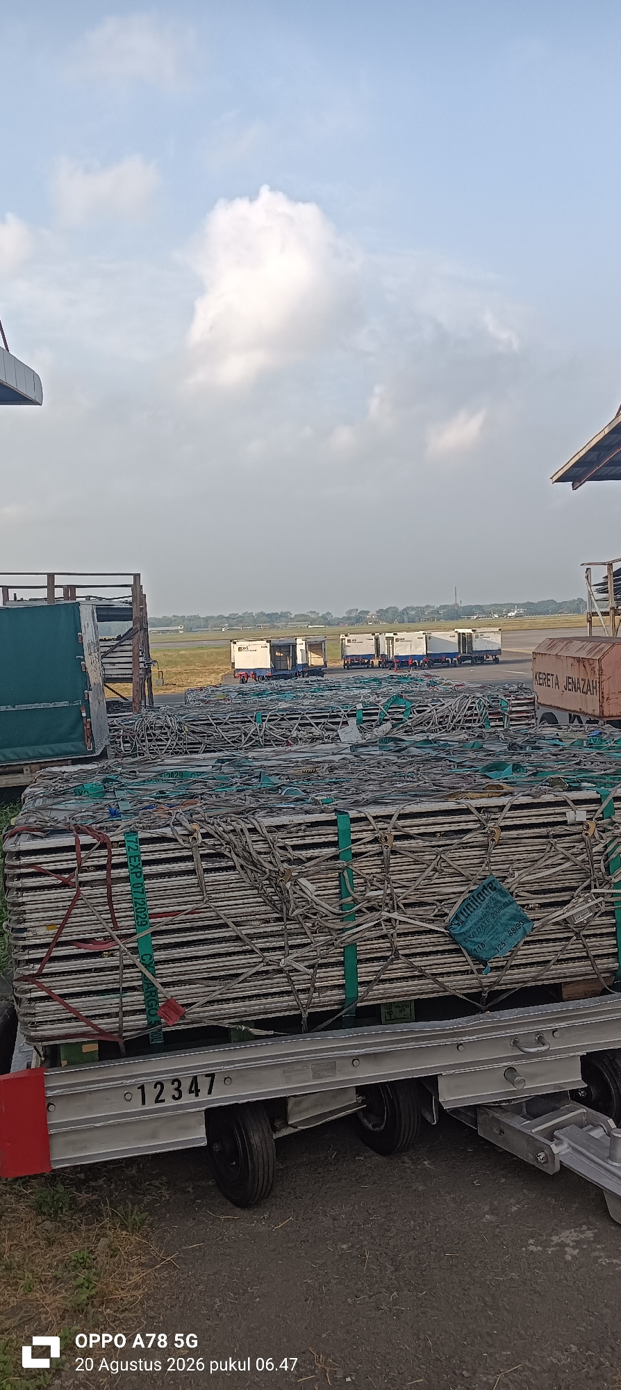 The image captures an outdoor scene at an airport, likely within the Sedati Agung area of Indonesia, given the timestamp on the photo which includes "Agustus" (August) and "pukul" (time in Indonesian). In the foreground, a large, flatbed trailer is loaded with multiple stacked rows of what appear to be aluminum or metal frames, possibly for scaffolding or construction. These stacks are secured with green straps and an intricate network of white ropes and netting. A serial number, "12347," is clearly visible on the side of the trailer.
In the mid-ground, several white cargo dollies or small trucks are lined up on the tarmac, some displaying "JAS" logos, suggesting a logistics or airline service company. These dollies are likely used for transporting luggage or cargo to and from aircraft. Beyond the dollies, a runway is visible stretching into the distance, with a hazy horizon and some scattered trees and buildings. The sky is partly cloudy, with large, soft white clouds against a pale blue backdrop.
To the right side of the image, part of a covered structure, possibly a hangar or a loading bay, is visible. A portion of a large vehicle, labeled "KERETA JENAZAH" (meaning "funeral car" in Indonesian), is also visible on the right, indicating a potential service for transporting deceased individuals, which might be related to the airport's operations or services. The ground is a mix of paved asphalt and some dry grass and dirt. The overall impression is one of an active, operational airport environment.