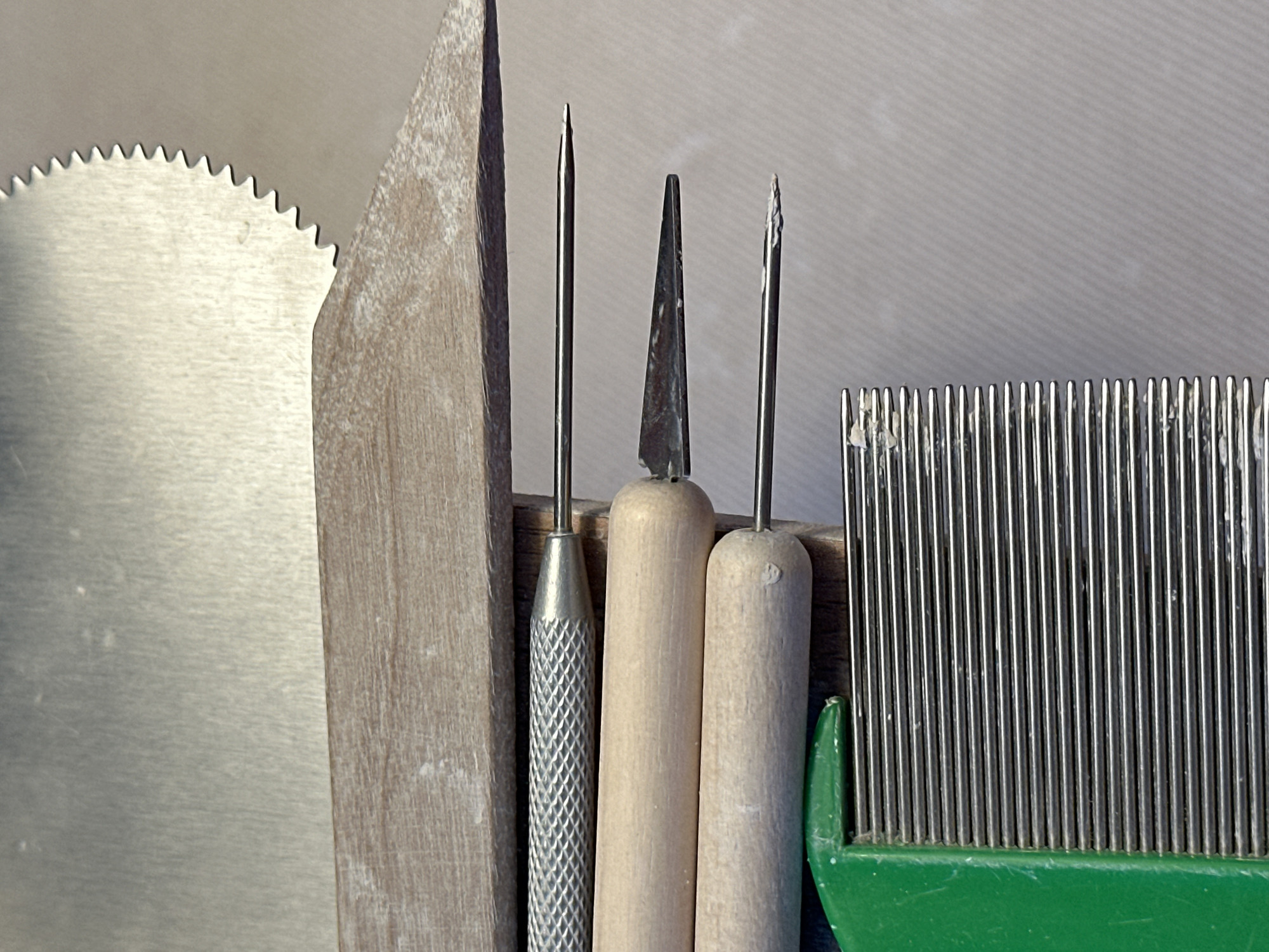 The image displays a close-up, overhead view of a collection of sculpting or pottery tools arranged on a textured, light-colored surface. In the foreground, a green plastic-handled tool with numerous thin metal tines, resembling a comb or rake, is visible on the left. Adjacent to and above this tool are several sculpting instruments. These include a pointed metal needle tool with a textured silver handle, two wooden-handled tools, one with a pointed metal end and another with a flat, triangular metal blade. Above these, a longer, flat wooden tool is positioned horizontally. At the very top of the image, a metallic tool with a serrated edge is partially visible, suggesting it is a rasp or saw. There are no people or any indications of the specific location within Nevinnomyssk, Russia. The overall impression is one of an organized workspace for crafting or artistic creation.