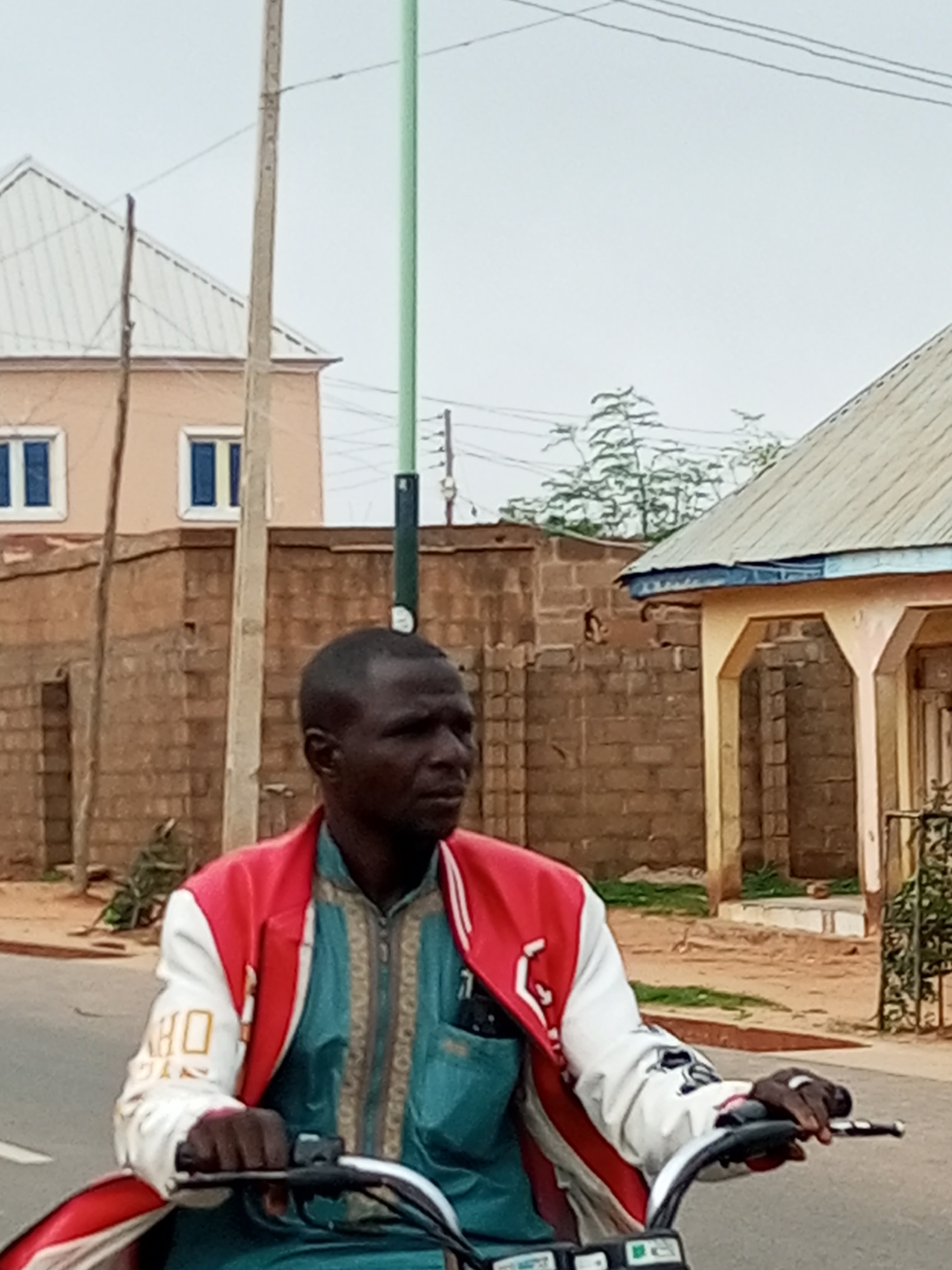 A man rides a motorcycle on a dirt road. He is wearing a red and white jacket over a teal garment. In the background, there are brick buildings and a light blue sky. A utility pole with wires stands to the left of the man. The scene is likely in Tempure, Nigeria, given the context.