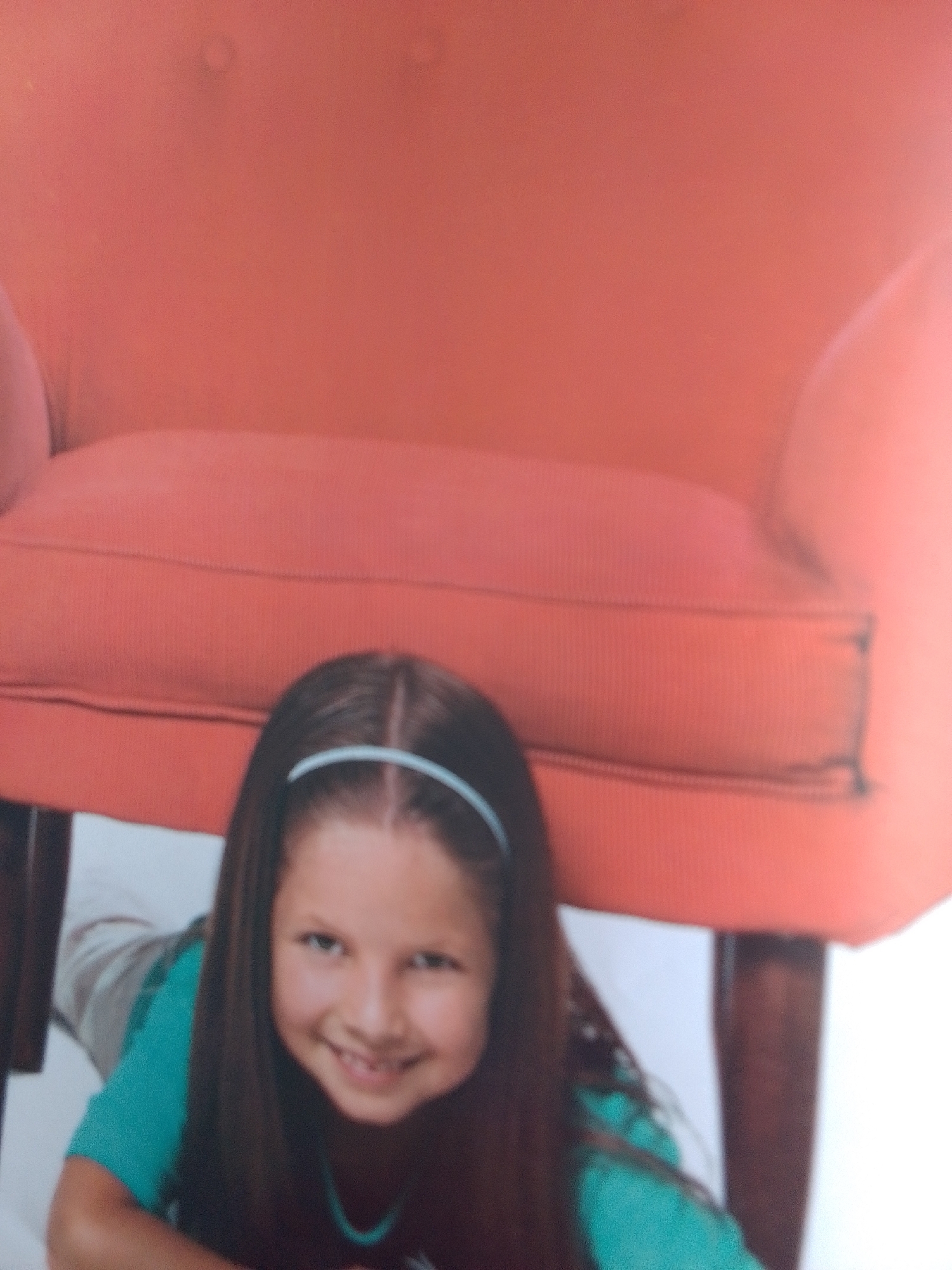 A young girl with dark hair and a blue headband lies on the floor in front of a large, coral-colored armchair. She is wearing a turquoise shirt and appears to be smiling at the camera. The background is a plain white wall. The image is blurry and lacks detail due to low resolution. There is no identifiable information about the location in La Tagua, Venezuela.