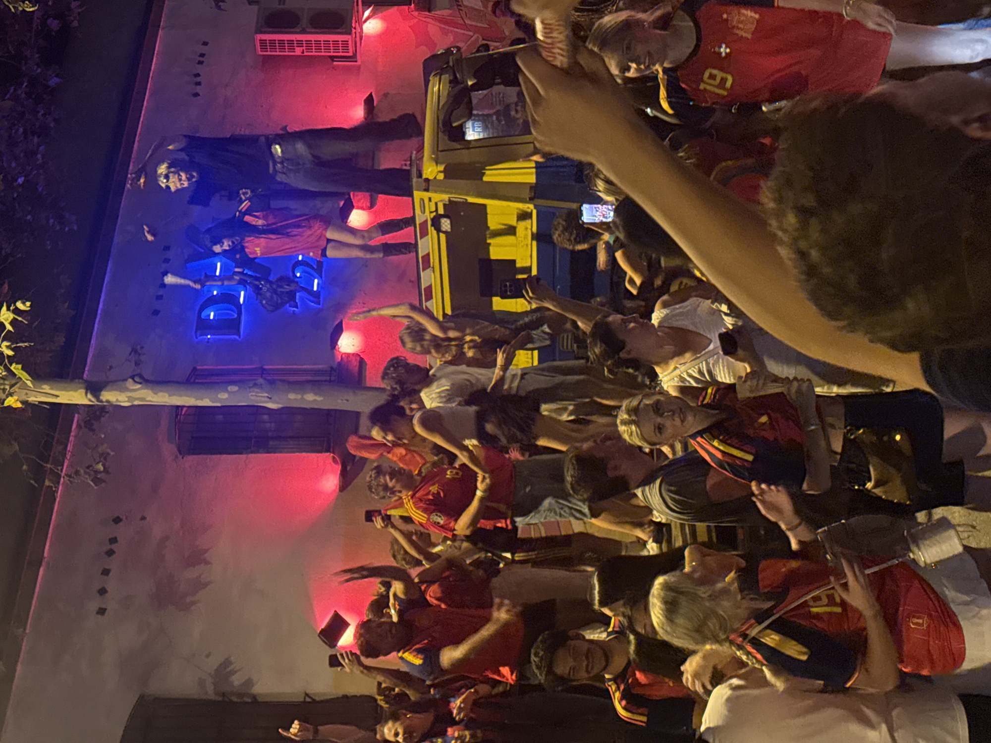 This image captures a lively street scene in Barcelona, Spain, likely during a celebration or event, given the energetic atmosphere. A crowd of people, many dressed in red and yellow attire suggesting support for a team or national identity, are gathered together. Some individuals are holding up their phones, possibly to record or photograph the events. In the background, a building with neon blue signage that reads "DI 5722" is illuminated by vibrant red and blue lights, contributing to the festive mood. A yellow and blue truck is parked on the street, and some people are standing on top of it, further enhancing the sense of celebration. The overall impression is one of a public gathering, filled with excitement and communal spirit, taking place at night in a Spanish urban setting.