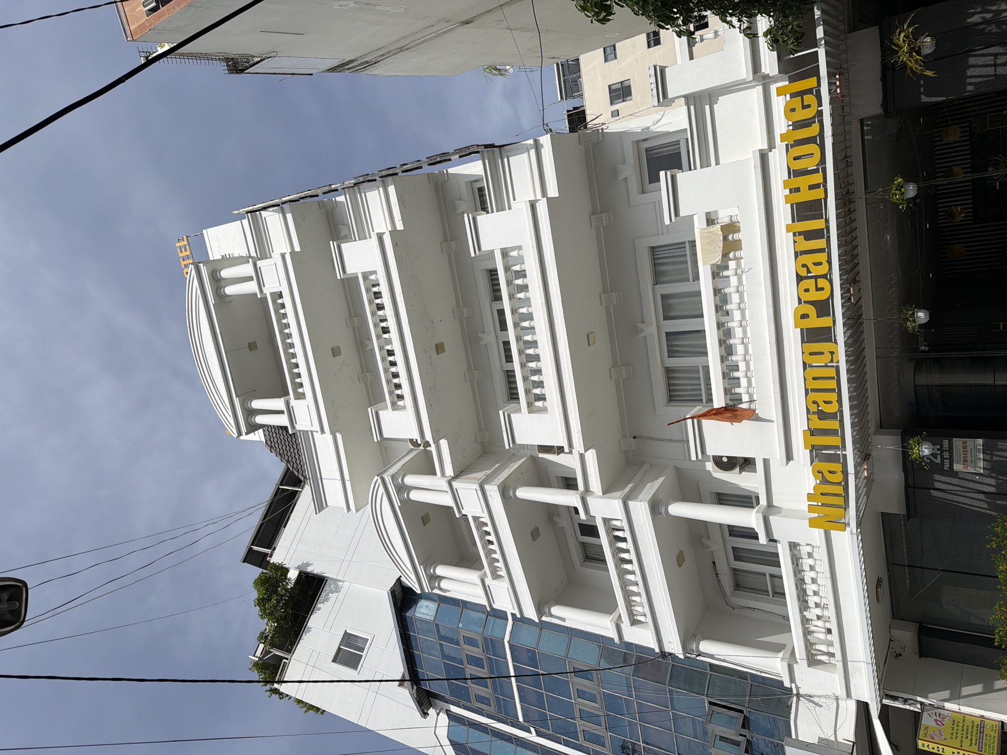 A white hotel building with multiple balconies, columns, and decorative elements stands under a cloudy sky in Nha Trang, Vietnam. The building has a prominent yellow sign that reads "Nha Trang Pearl Hotel". A modern building with a blue glass facade is visible to the left of the hotel. The foreground shows the entrance to the hotel, with large windows and some hanging plants. A small orange flag is attached to the hotel. Overhead power lines are visible against the sky.