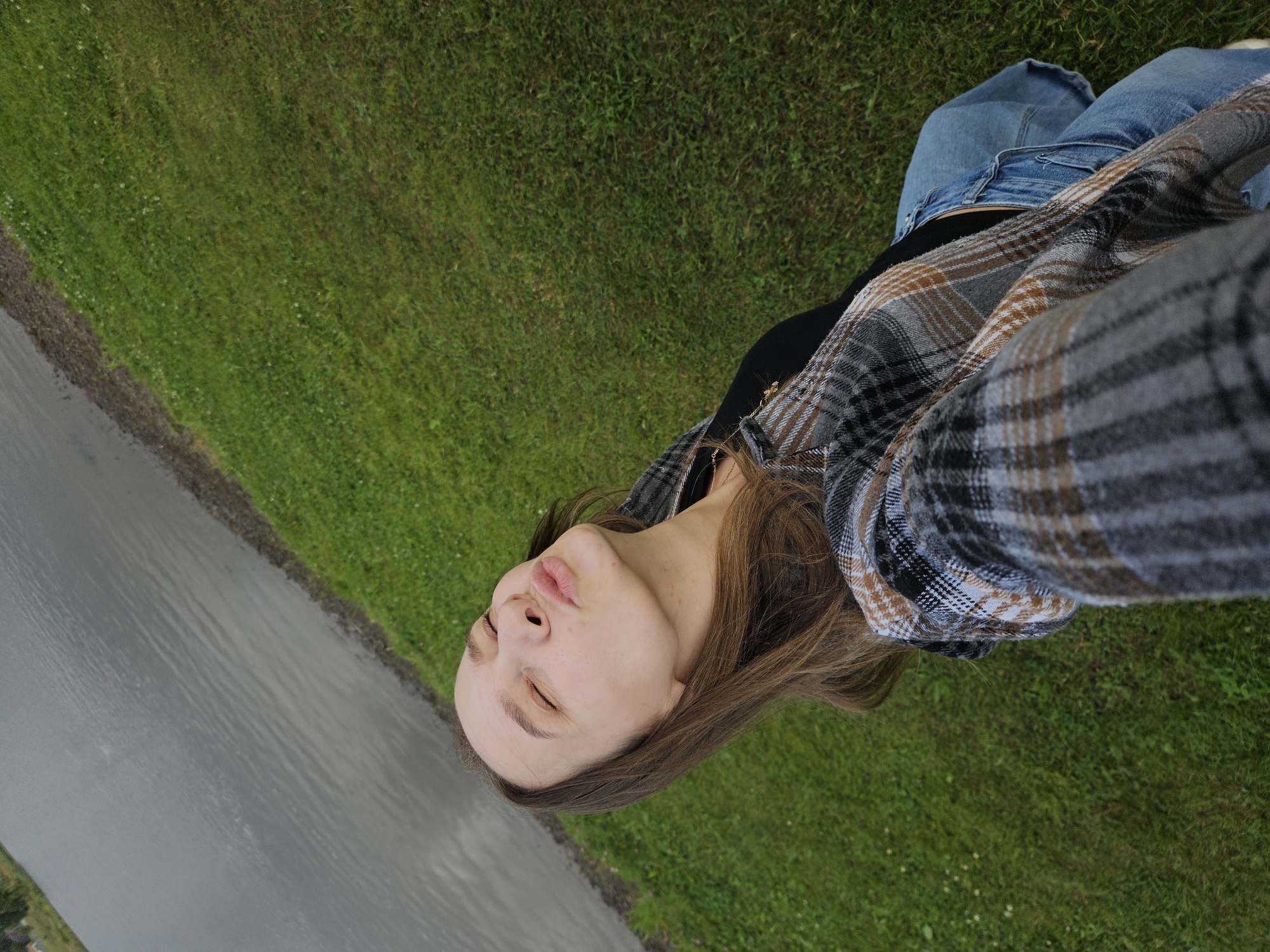 A young woman with brown hair is taking a selfie outdoors. She is wearing a plaid shirt and jeans. Her eyes are closed, and her lips are puckered as if she is about to kiss. She is standing on a grassy area next to a body of water. The environment suggests a park or recreational area, possibly near the Kama River in Elovo, Russia, given the proximity to water and open green space. The lighting is overcast, indicating a cloudy day.