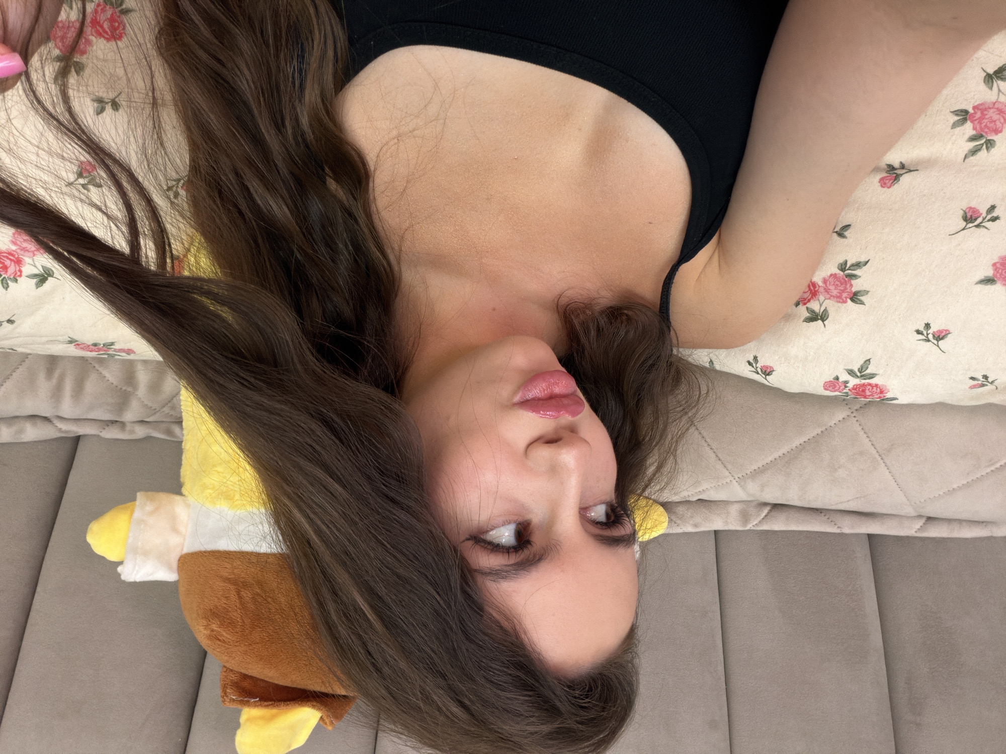 The image shows a young woman with long, dark brown hair and fair skin, wearing a black tank top. She is lying down on a bed with a floral patterned pillow and a beige headboard. A stuffed animal, mostly yellow with brown and white accents, is visible behind her. She is looking to her left and her lips are slightly parted. The scene appears to be indoors, likely in a bedroom. The image does not contain any elements that would indicate a specific location in Chaikovsky, Russia.