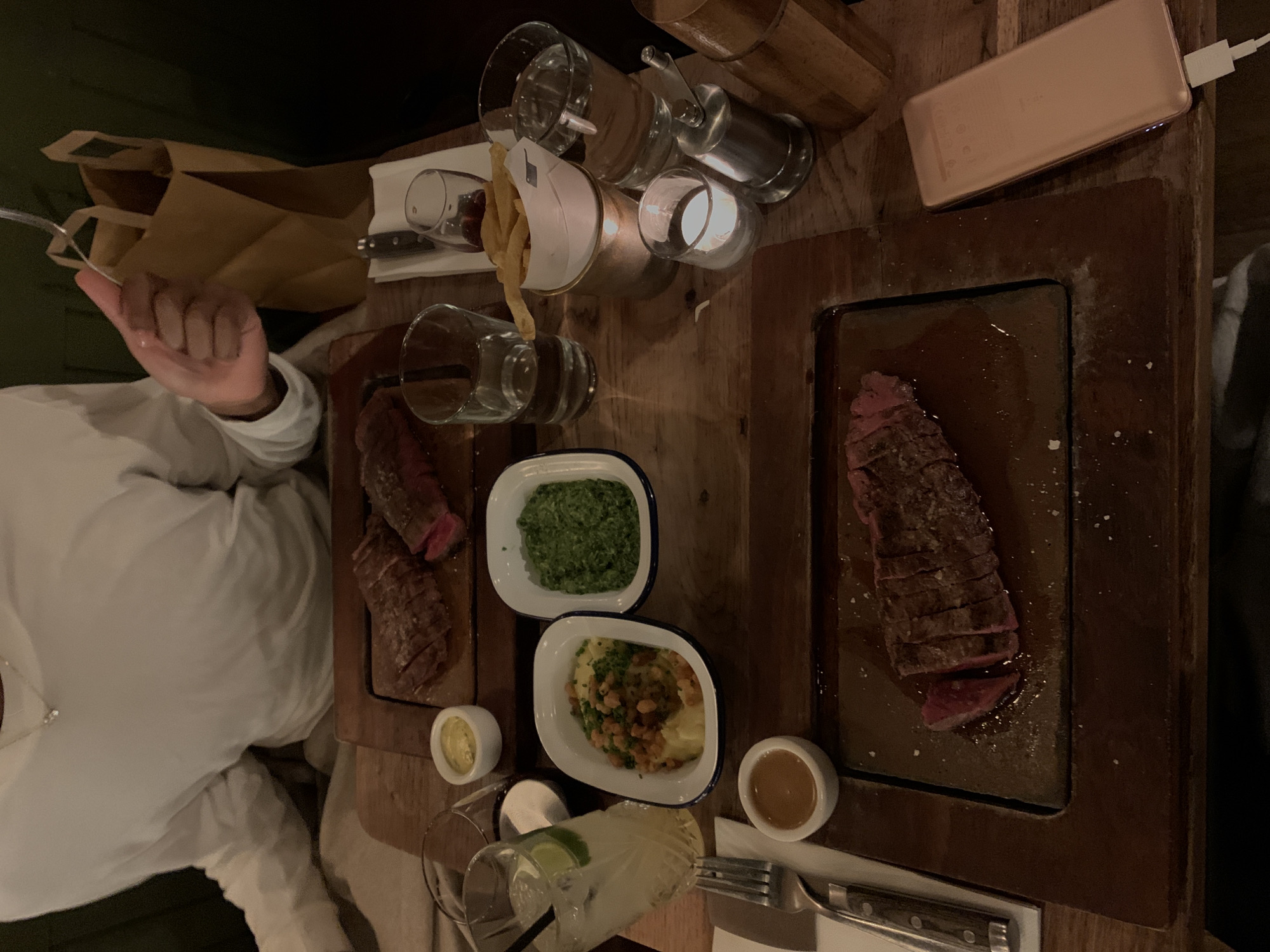 The image displays a dimly lit dinner scene at a restaurant, captured from an overhead perspective, likely during the evening. A wooden table is laden with food and drink, creating a warm and intimate ambiance.
One person is partially visible on the left side of the frame, wearing a white long-sleeved shirt. Their hand is holding a silver fork, poised as if about to eat.
The table features two main dishes of sliced, cooked meat, likely steak, presented on dark wooden boards. The meat appears to be cooked to a medium-rare or medium doneness, with pink centers and sprinkled with seasoning. Accompanying the meat are two white enamel dishes with blue rims, one filled with green creamed spinach or similar greens, and the other with mashed potatoes garnished with chives and crispy toppings. Small white ramekins hold a yellowish sauce or butter and a brown gravy or jus. A bowl of golden french fries is also present.
For drinks, several clear glasses are visible. One contains a clear cocktail with ice and a lime wedge, while others appear to hold water or other beverages. A smaller glass with red liquid, possibly wine, is also on the table.
Other notable objects include a rose gold portable power bank connected by a white cable, resting on the wooden table. A small candle in a glass holder provides additional soft light. In the upper left background, brown paper bags are stacked. Cutlery, including forks with dark handles, and a white napkin complete the setting. The general atmosphere suggests a relaxed and enjoyable dining experience in a restaurant setting, consistent with establishments found in London. No clear, readable text is visible on any objects, though there are blurred markings on the power bank.