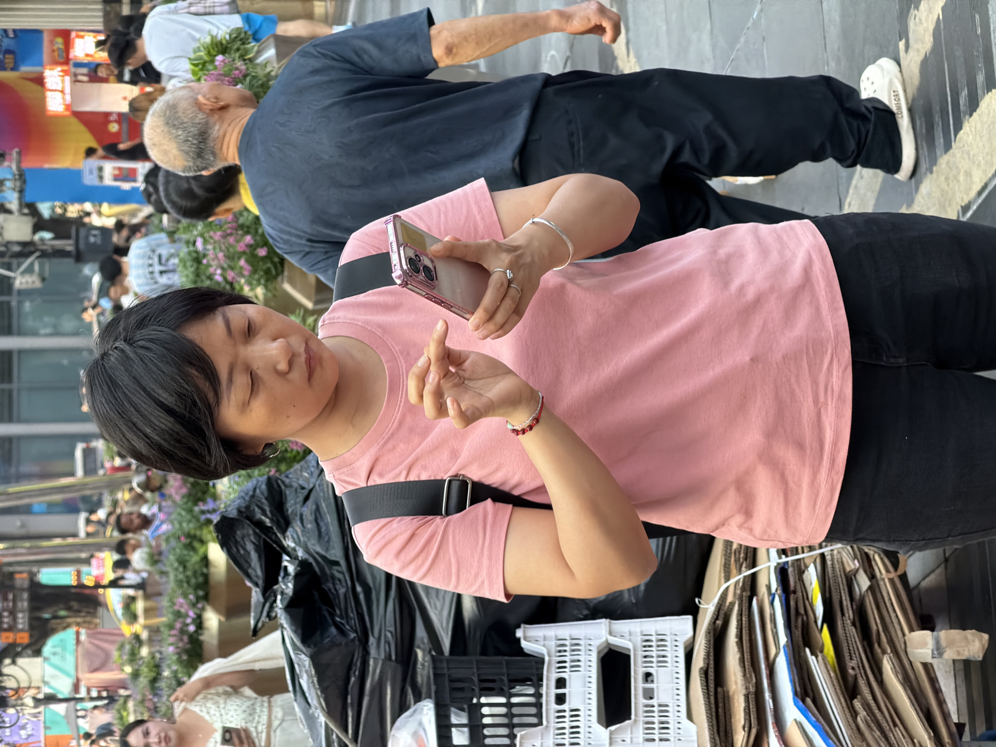 In Yuzhong District, China, a woman in a pink t-shirt and black pants stands focused on her smartphone. She wears a silver bracelet on her right wrist and rings on her left hand. Behind her, a black garbage bag is partially visible, next to stacked white plastic crates and a pile of cardboard. Further in the background, other people are walking, and the scene appears to be a busy urban street with colorful signage and landscaping.